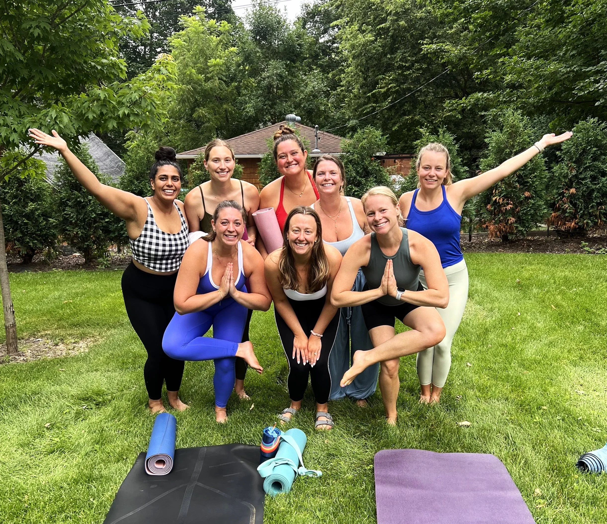 Group of smiling women in athletic clothing practicing yoga outdoors on a lawn, with yoga mats and fitness equipment on the ground, surrounded by lush green trees and shrubs.