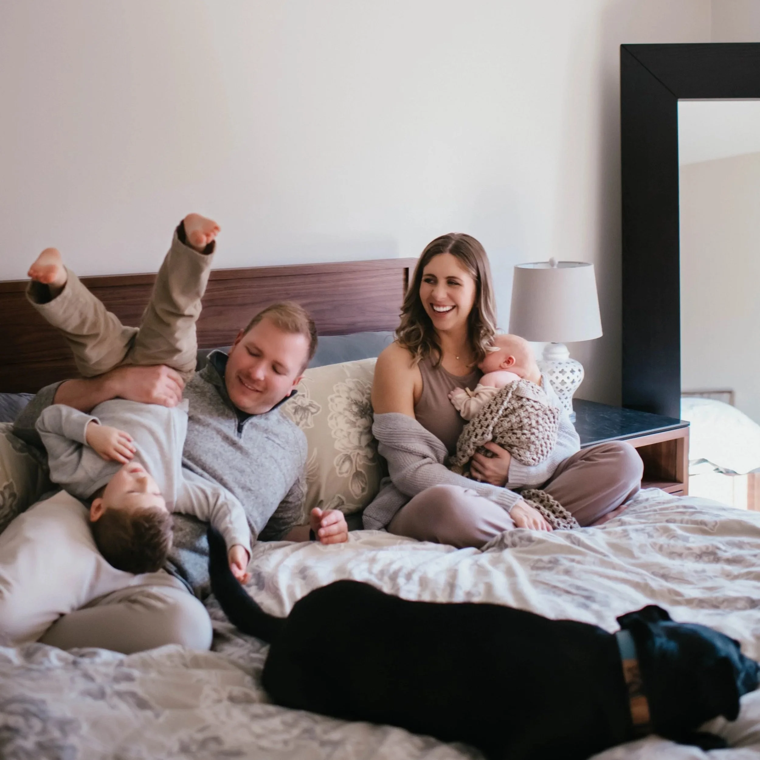 A family relaxing and playing on the bed in a cozy room, with a young woman smiling, a man, and children including a baby. The bed has a textured white blanket, and a nightstand with a lamp is visible. A large black-framed mirror hangs on the wall.