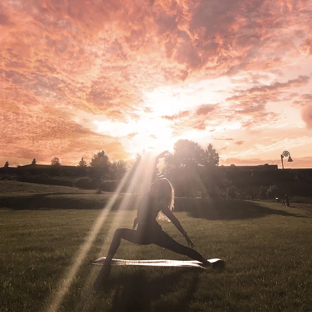 A person practicing yoga outdoors at sunset, silhouetted against a colorful sky with clouds. The individual is in a low lunge pose on a mat in a grassy field, with the sun shining through trees in the background.