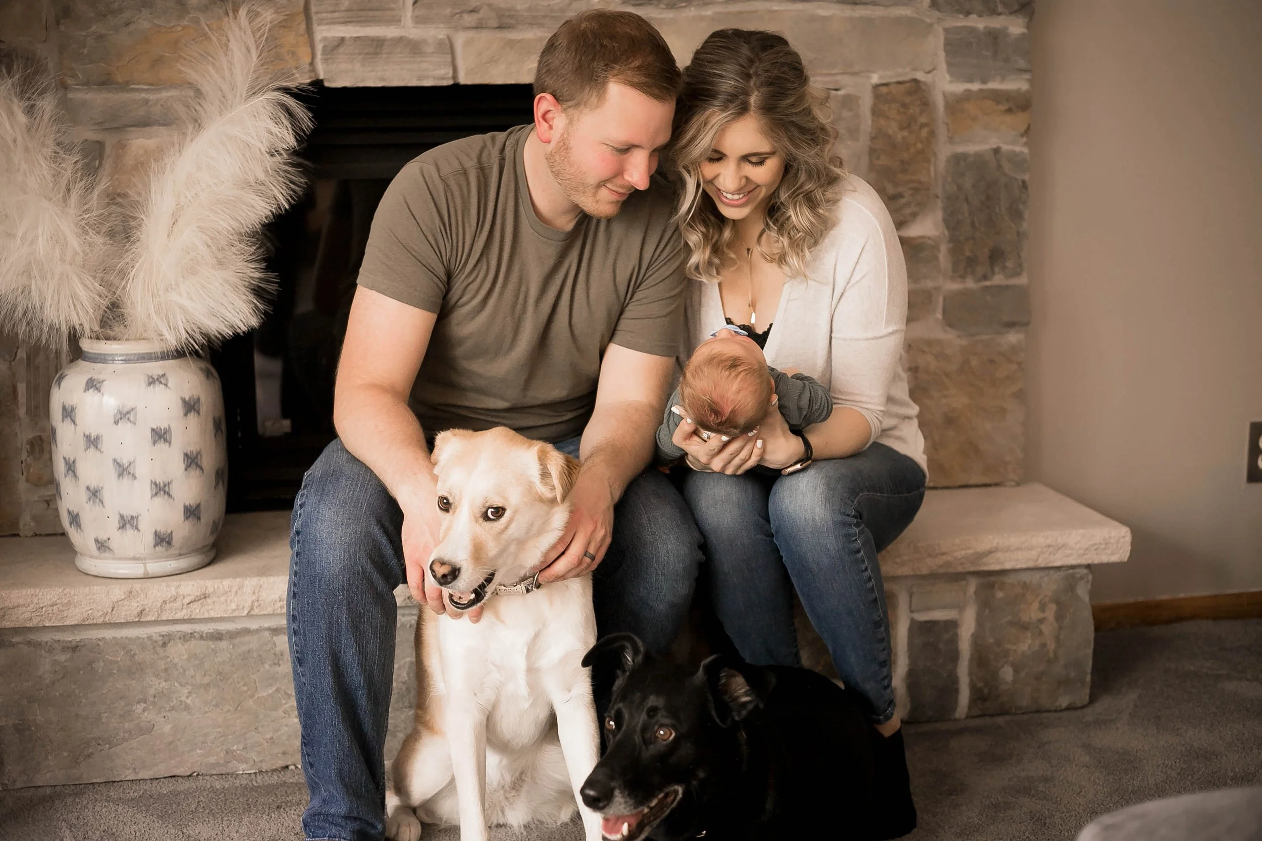 A couple sitting on a stone fireplace hearth with two dogs and a baby. The man is holding one dog, and the woman is holding the baby, all smiling and looking at each other. There is a large decorative vase with pampas grass next to them.
