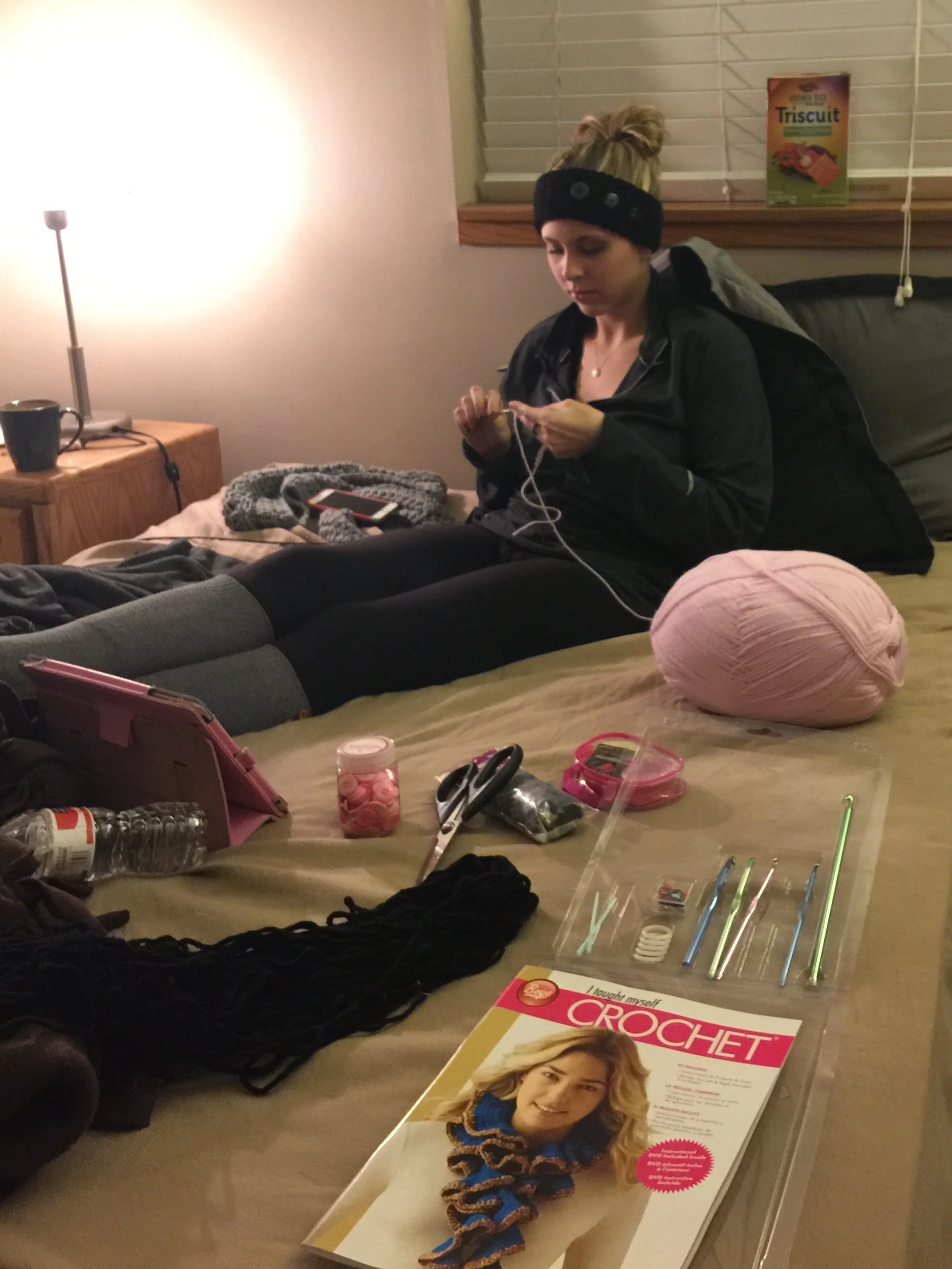 A young woman with blonde hair tied in a bun, wearing a black headband and black jacket, sits on a couch looking at her phone, surrounded by yarn, knitting needles, and a knitting pattern magazine on the table.