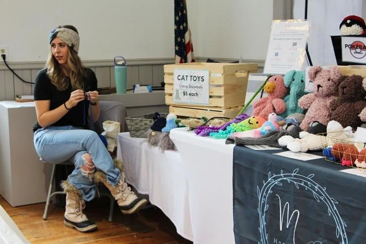 A woman sitting at a table with handmade knitted plush toys for sale, including cats and bears, at a craft fair. The table has signs and a black cloth with a logo.