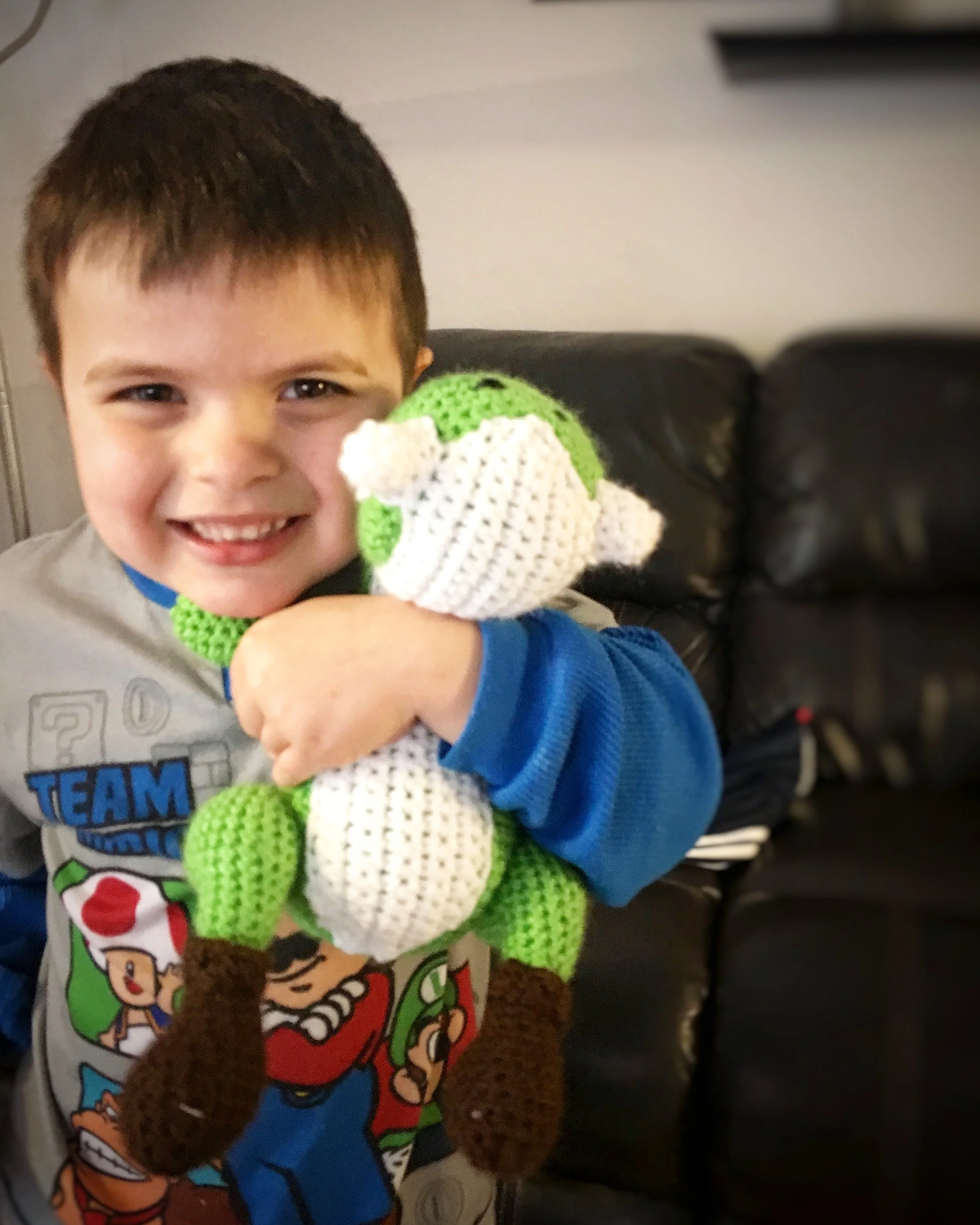 Young boy with dark hair smiling, holding a green and white crocheted puppet with brown limbs and white face, sitting on a black leather couch.