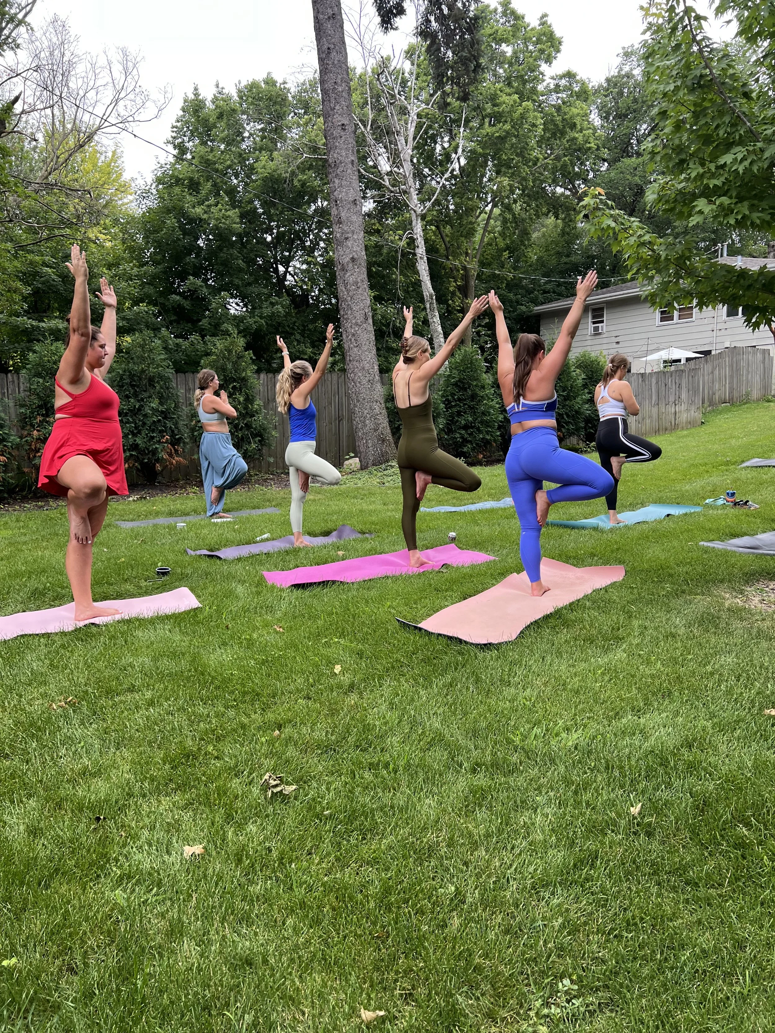 A group of women practicing yoga outdoors in a backyard, standing on yoga mats in tree pose with one leg bent and foot placed on the thigh of the opposite leg, arms raised overhead. They are dressed in workout clothing, and there are trees, a wooden fence, and a house in the background.