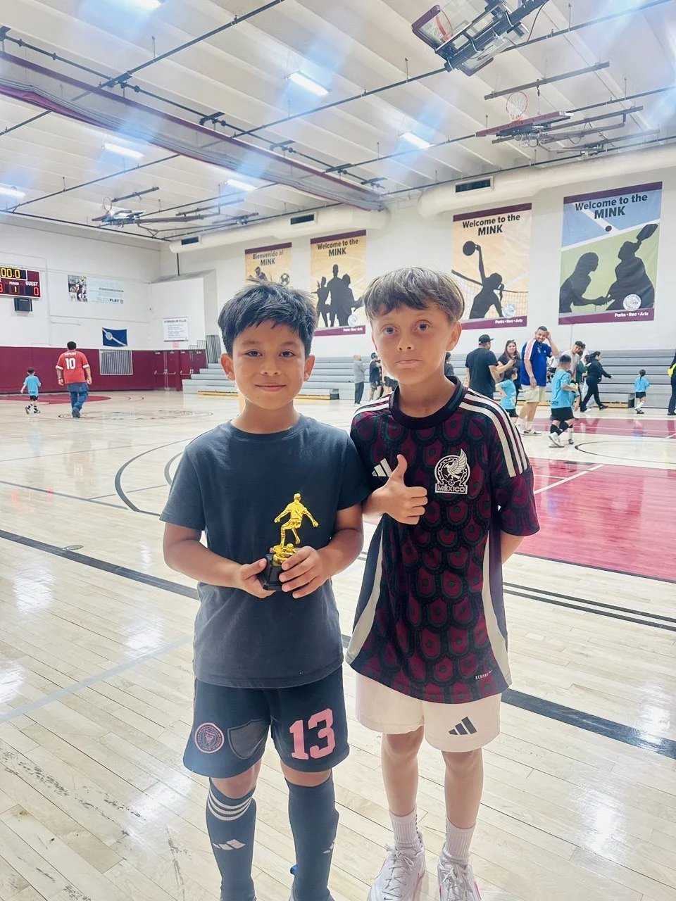 Two young boys standing in an indoor basketball gymnasium. The boy on the left is holding a small gold trophy, wearing a gray T-shirt and black shorts with pink and gray design, and sports knee-high black socks. The boy on the right is giving a thumbs-up, wearing a black and burgundy soccer shirt with white shorts. In the background, other children and adults are playing and walking, with banners and a scoreboard visible on the wall.