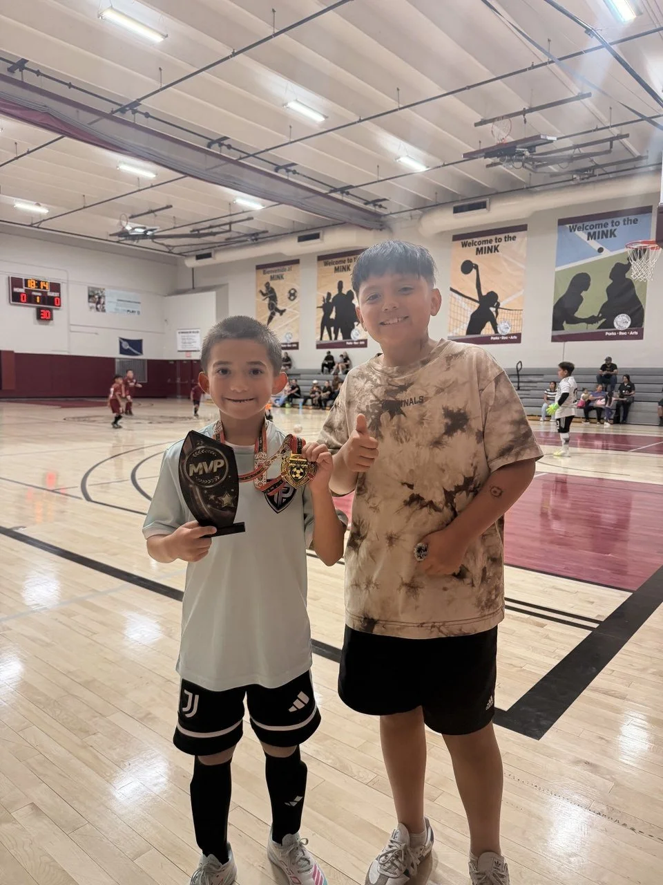 Two young boys standing on a basketball court inside a gymnasium, one holding an MVP award and medals, smiling at the camera. Other children are playing futsal in the background, with banners and a scoreboard in the local Vegas Recreational gym.