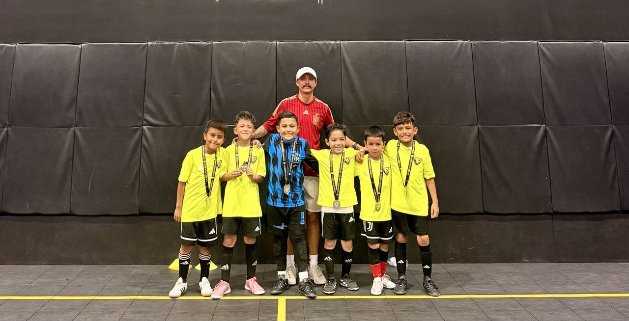 Six young boys wearing yellow soccer uniforms with medals around their necks and a coach in a red shirt standing behind them on an indoor futsal court during the Invictus Tournament.