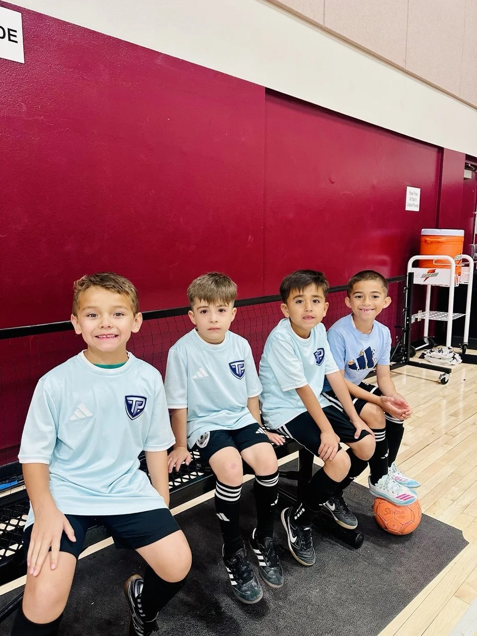 Four young boys in soccer uniforms sitting on a bench in a gym, with a soccer ball at their feet.