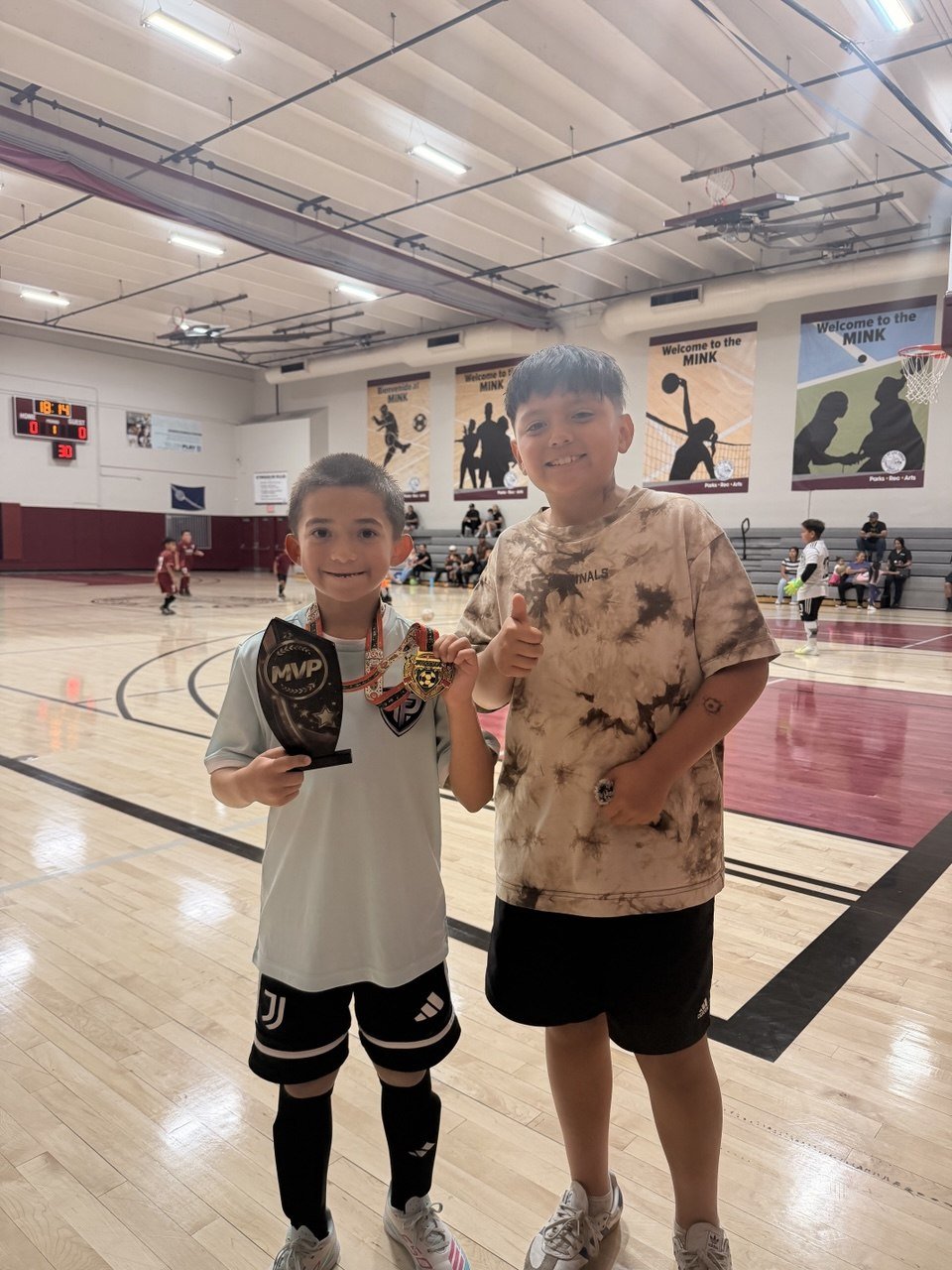Two boys at an indoor basketball court, one holding an MVP plaque and medals, smiling, with other children playing in the background and banners on the wall.