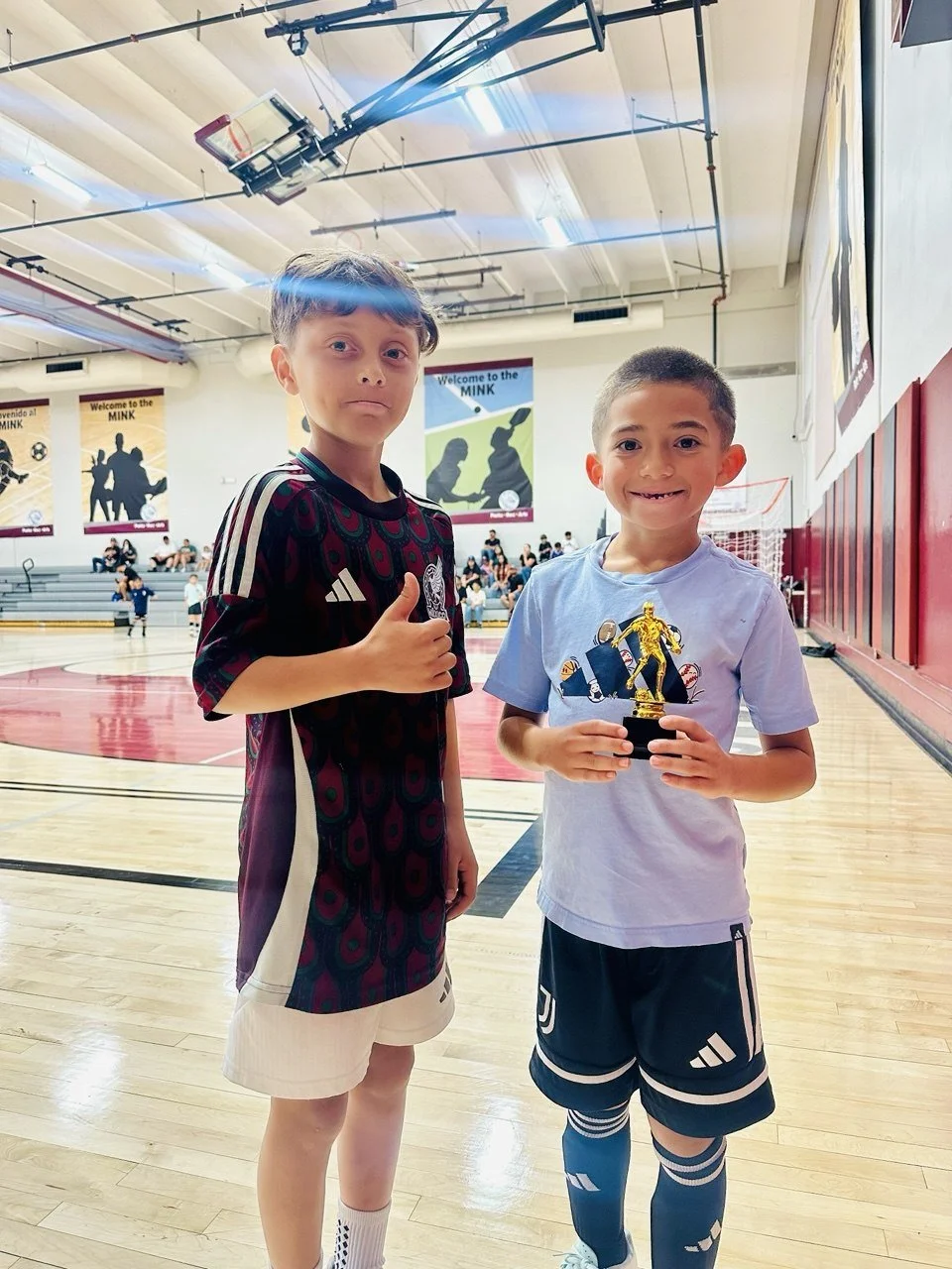 Two young boys standing on a basketball court inside a gymnasium. One boy is giving a thumbs-up and the other is holding a trophy. There are banners on the wall and people sitting on the bleachers in the background.