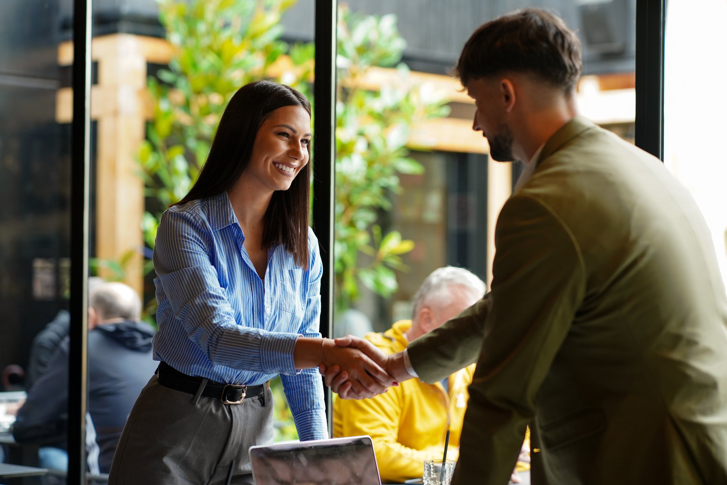 A woman in a blue striped shirt and gray pants shaking hands with a man in a beige jacket at a cafe or restaurant. They are smiling and engaging in a friendly conversation, with an outdoor setting and other people in the background.