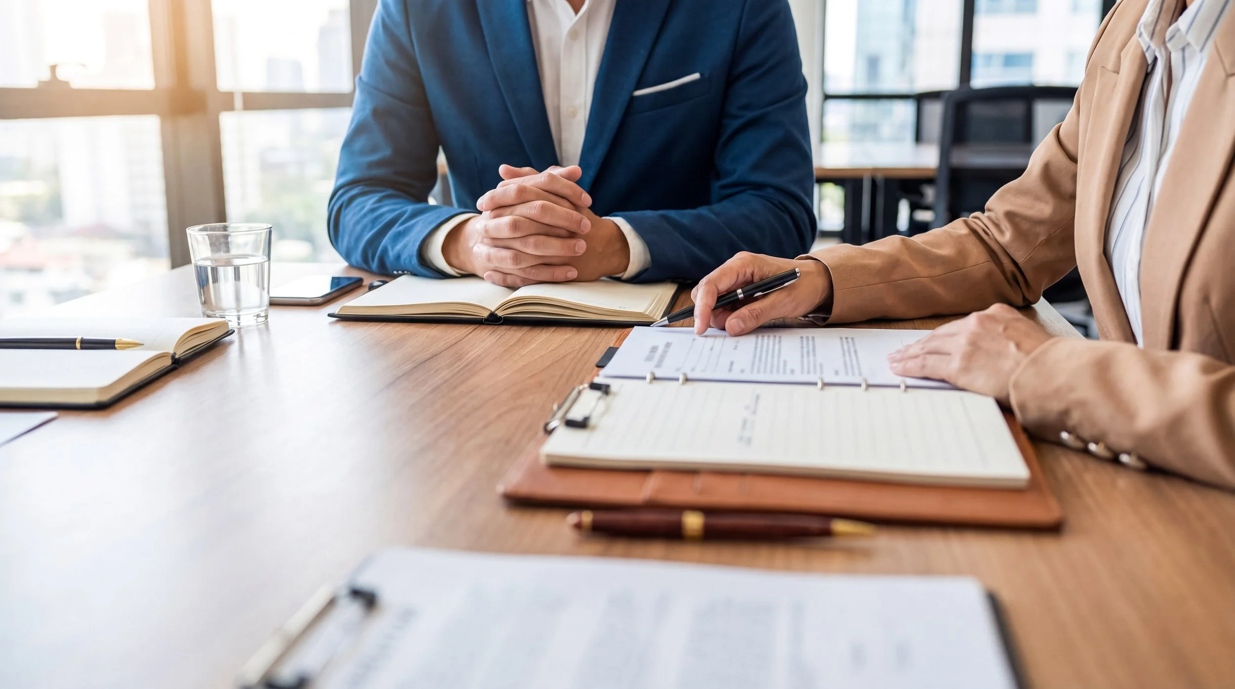 Two people in business attire sitting at a table during a meeting, with notebooks, documents, a tablet, and a glass of water on the table.