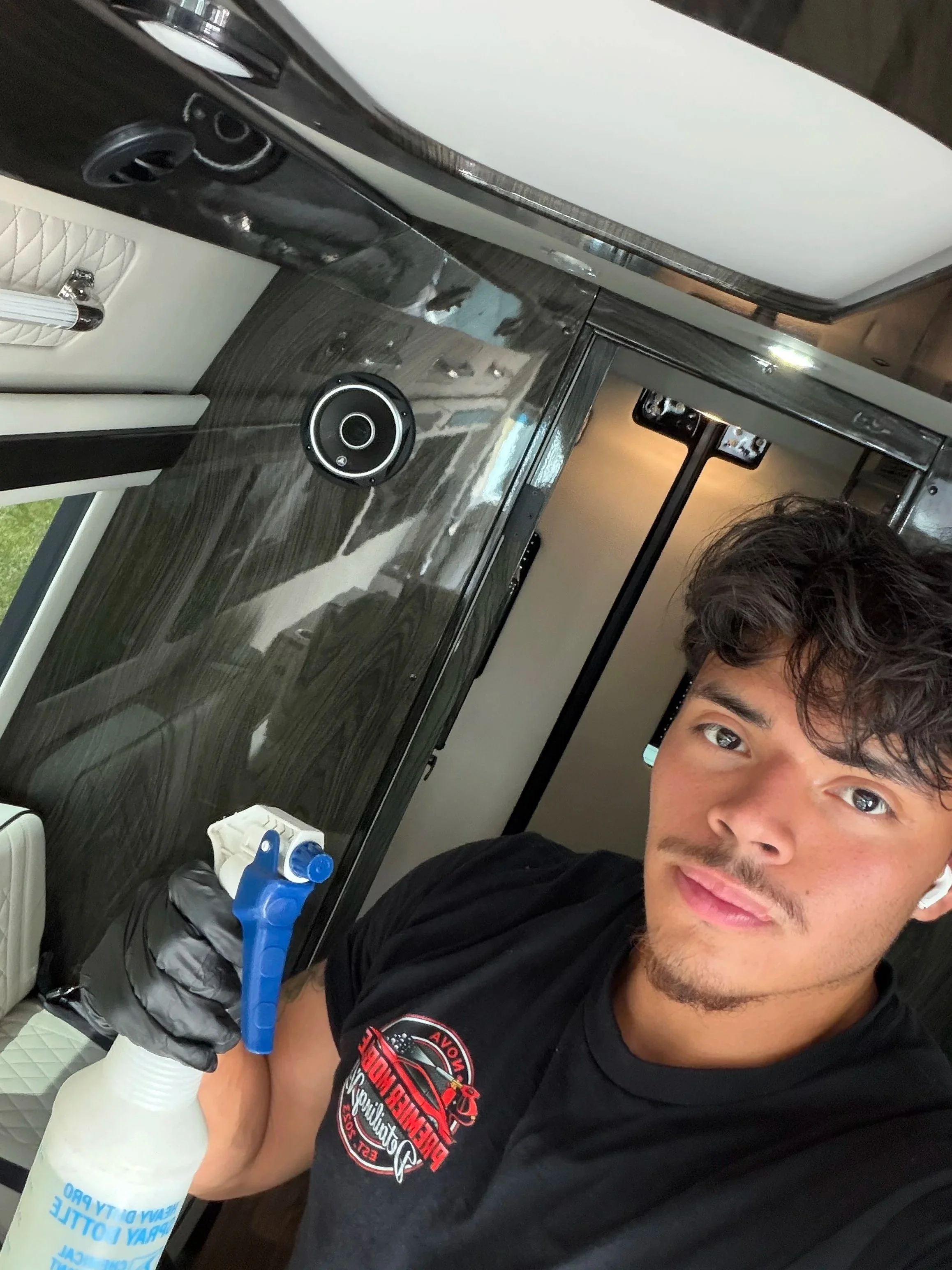 A young man with curly brown hair and a mustache taking a selfie during a hair donation event. He is holding a large plastic bottle of water and wearing a black T-shirt with a racing logo. The background shows a vehicle's interior with dark wood paneling and a speaker.