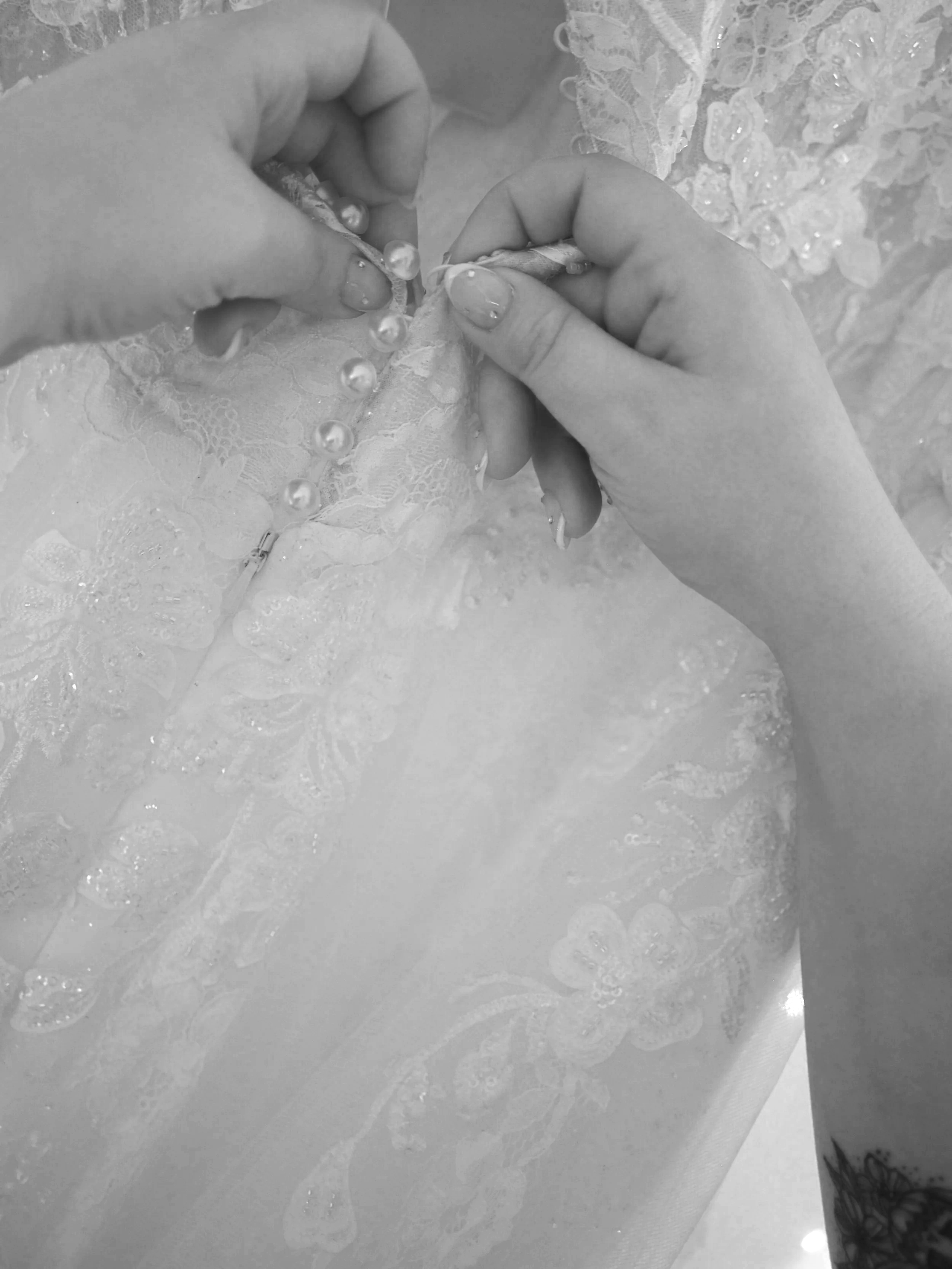 Close-up of hands stitching lace fabric with pearl and bead embellishments, with intricate lace and floral details visible.