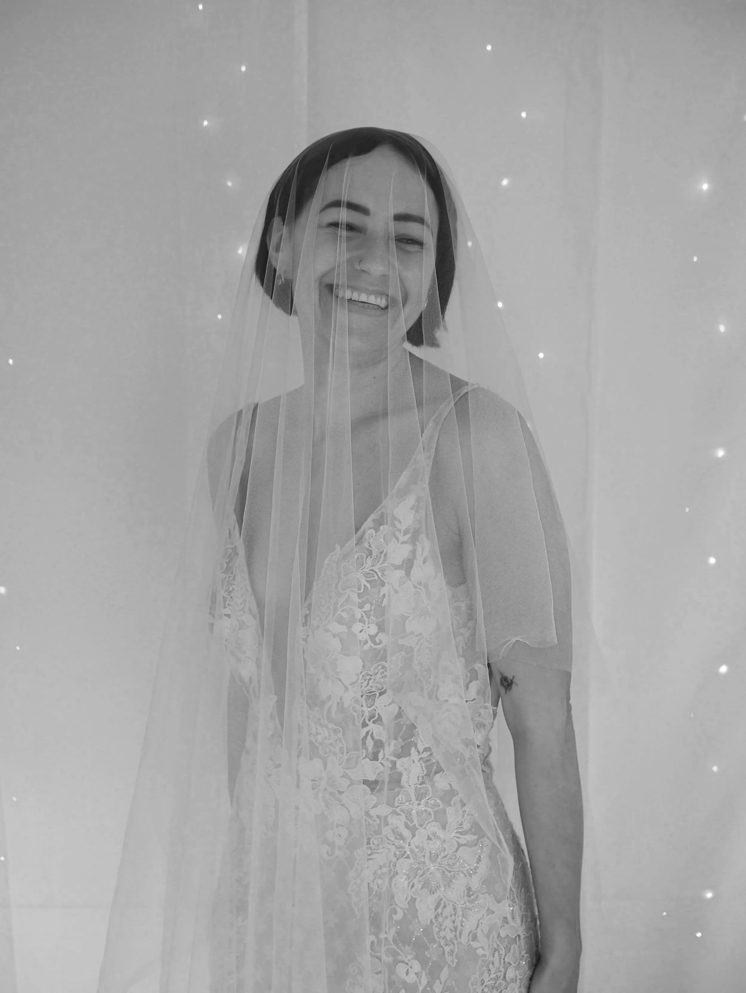 Black and white photo of woman in lace dress with a veil, smiling and standing against a background with small lights.