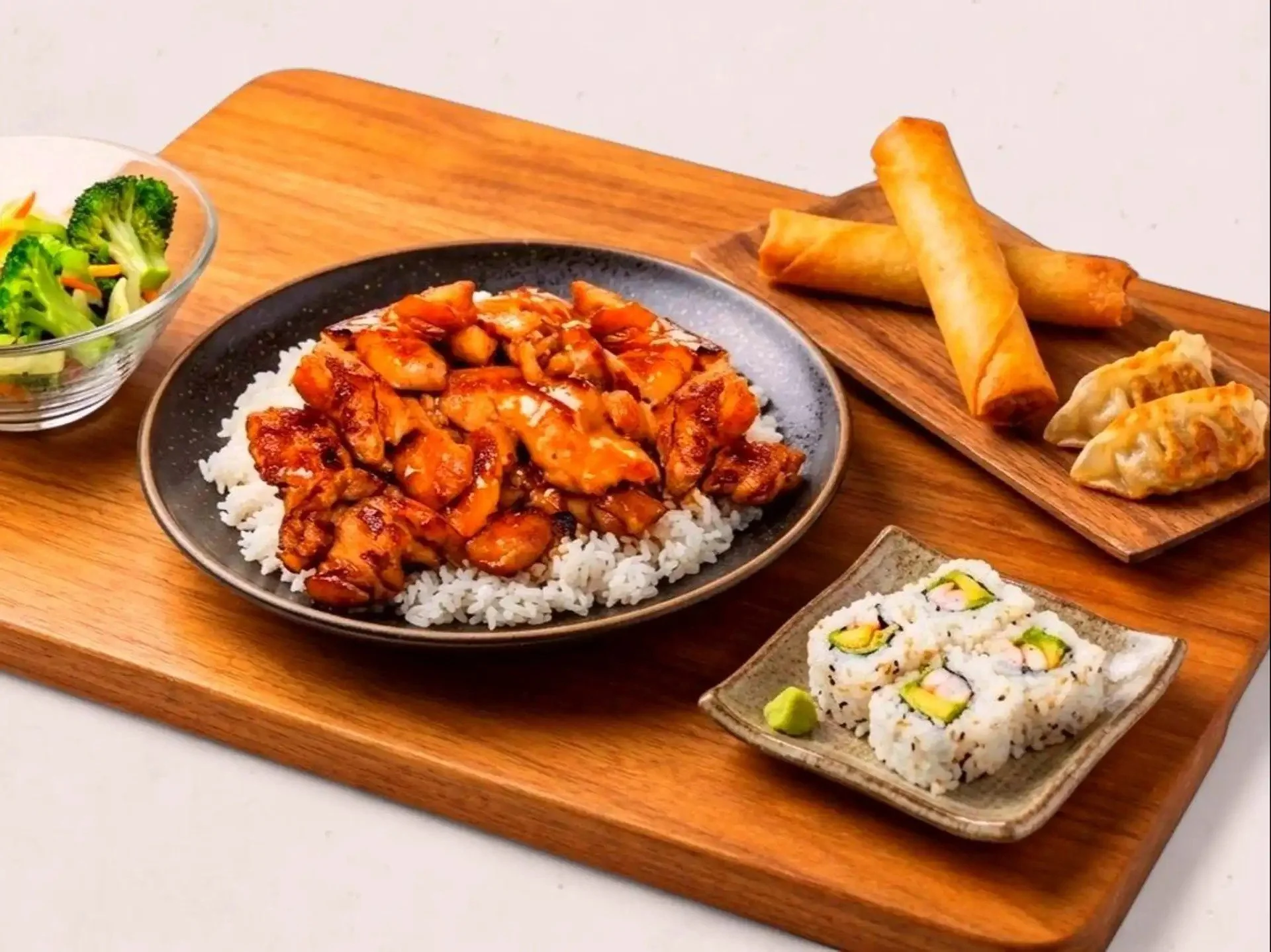 Assorted Japanese food dishes including a bowl of rice with stir-fried chicken and vegetables, a plate of spring rolls, dumplings, and sushi rolls with avocado, served on a wooden platter.