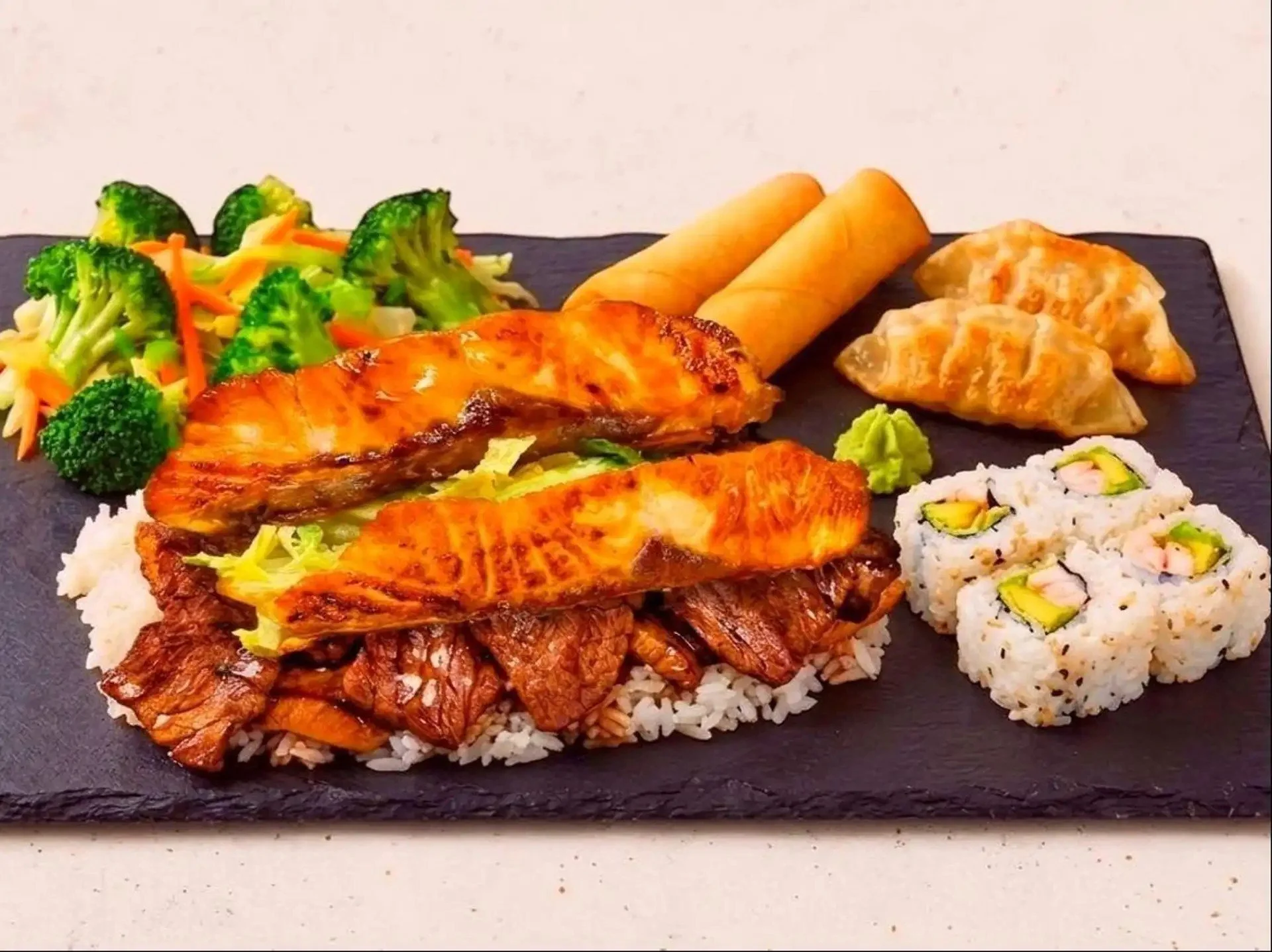 Assorted sushi, grilled salmon, Asian salad, spring rolls, and wasabi on a black slate serving platter.