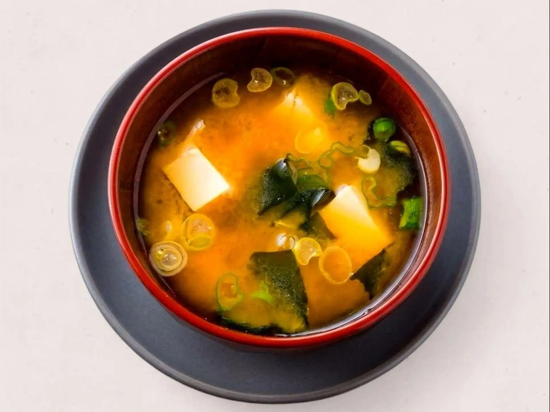 A bowl of miso soup with green onions, seaweed, and tofu on a black plate, viewed from above on a white surface.