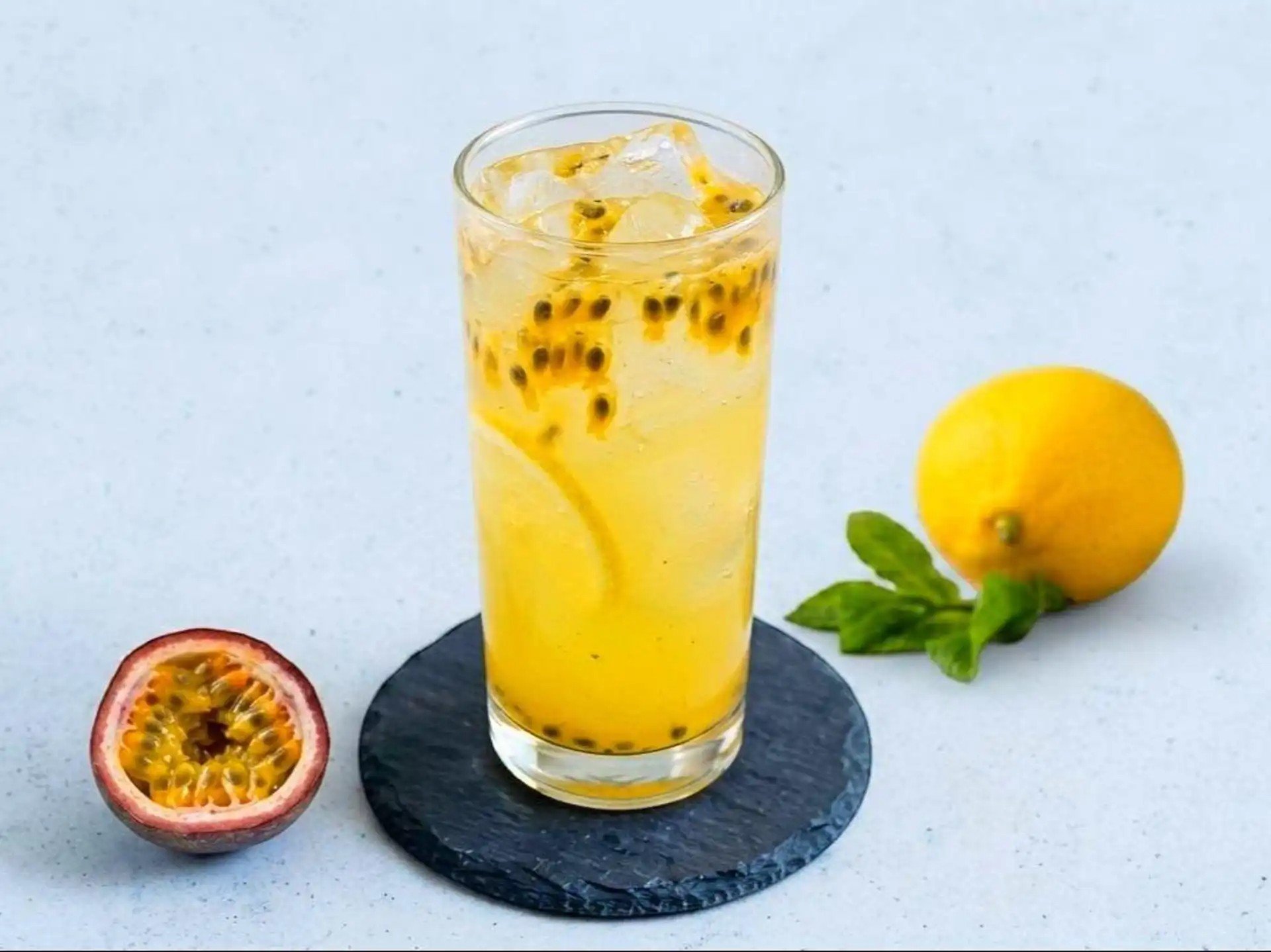 A tall glass of passion fruit juice with ice and passion fruit seeds, garnished with lemon slices, placed on a black slate coaster. Nearby, there is a sliced passion fruit, a whole lemon, and some fresh mint leaves.