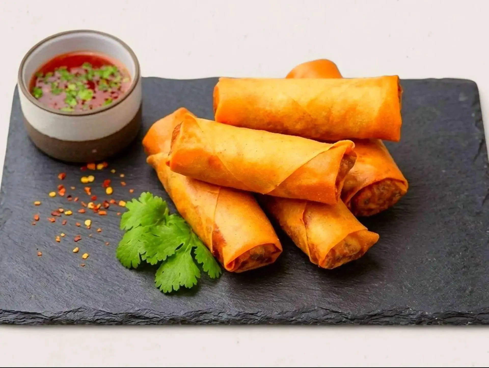 Four spring rolls on a black slate serving platter with a small bowl of dipping sauce and garnished with cilantro and red pepper flakes.