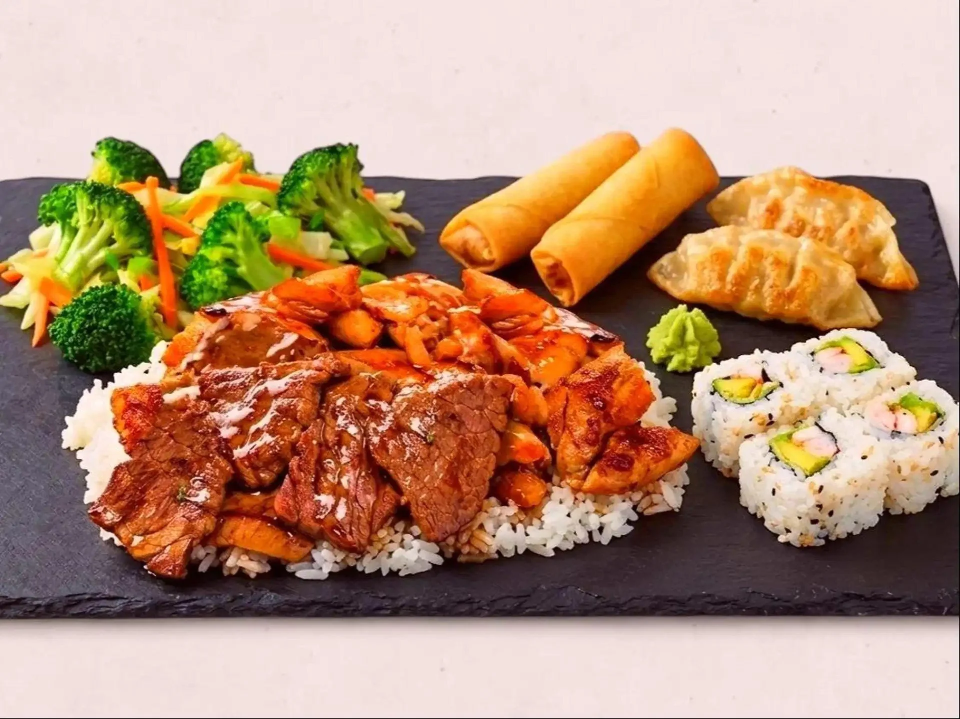 Sushi platter with broccoli, spring rolls, grilled beef, dumplings, and sushi rolls on a black slate plate.