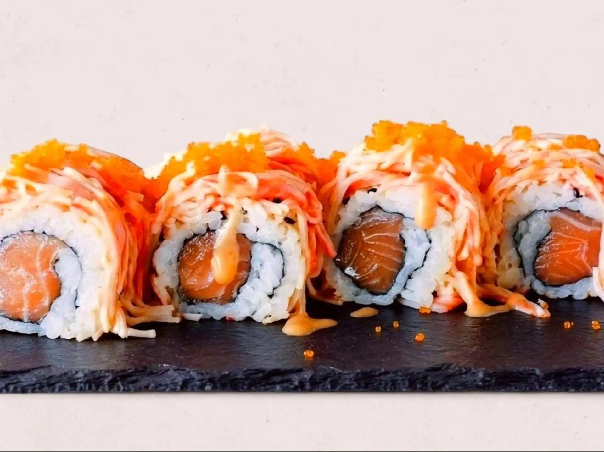 Close-up of sushi rolls with orange fish, cream cheese, and rice, topped with shredded crab meat and drizzled with spicy orange sauce on a black slate platter.