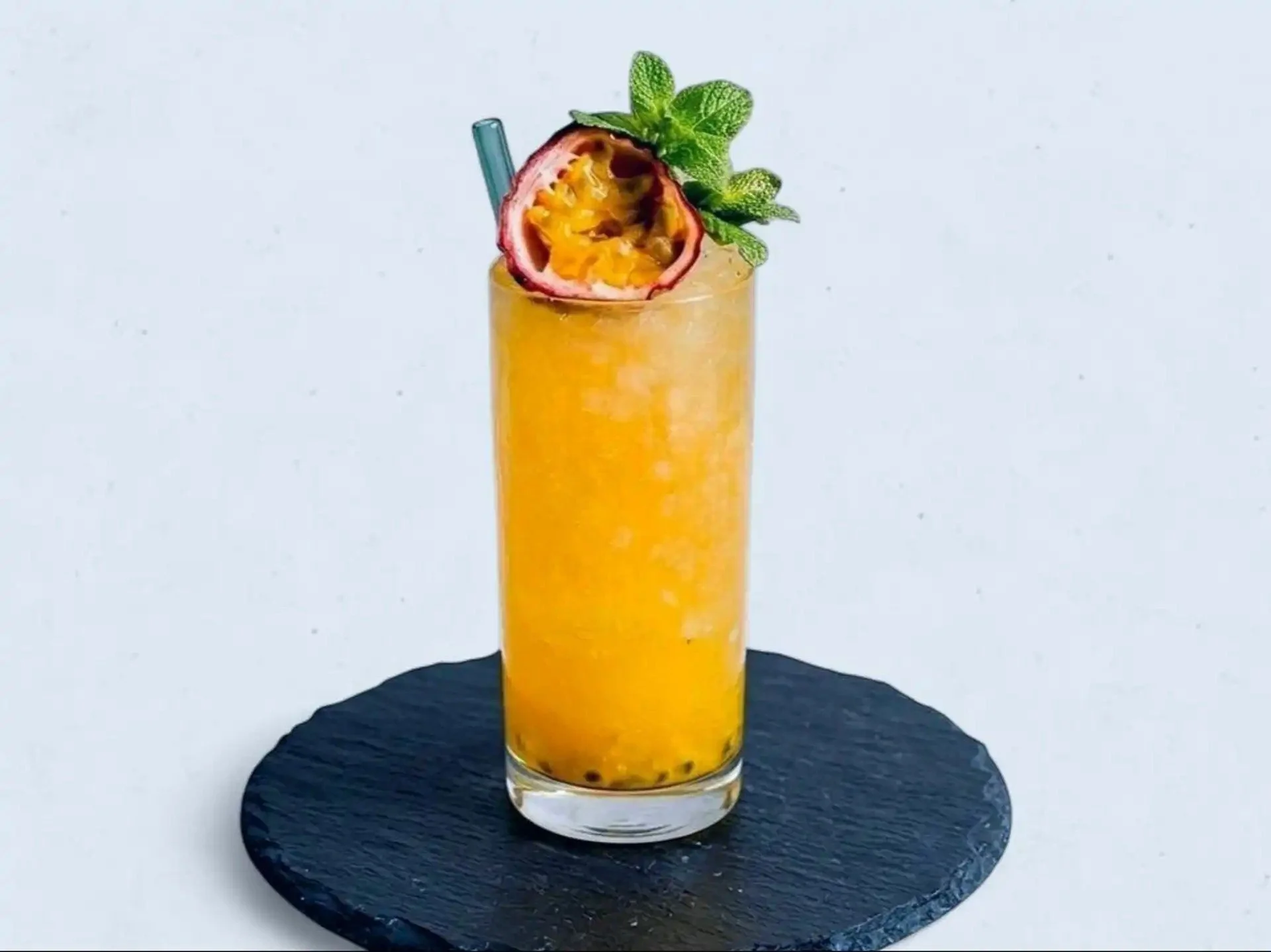 A tall glass of passion fruit and mango smoothie garnished with a halved passion fruit and a sprig of mint, sitting on a black slate coaster on a white surface.