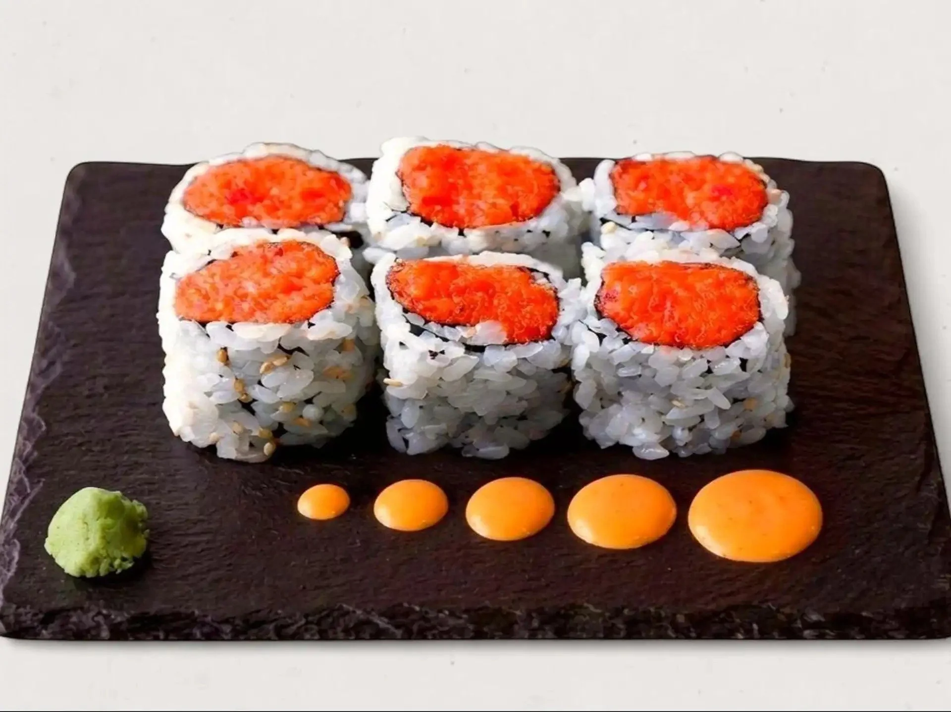 Six sushi rolls with orange fish roe, accompanied by wasabi, pickled ginger, and orange candies on a black rectangular plate.