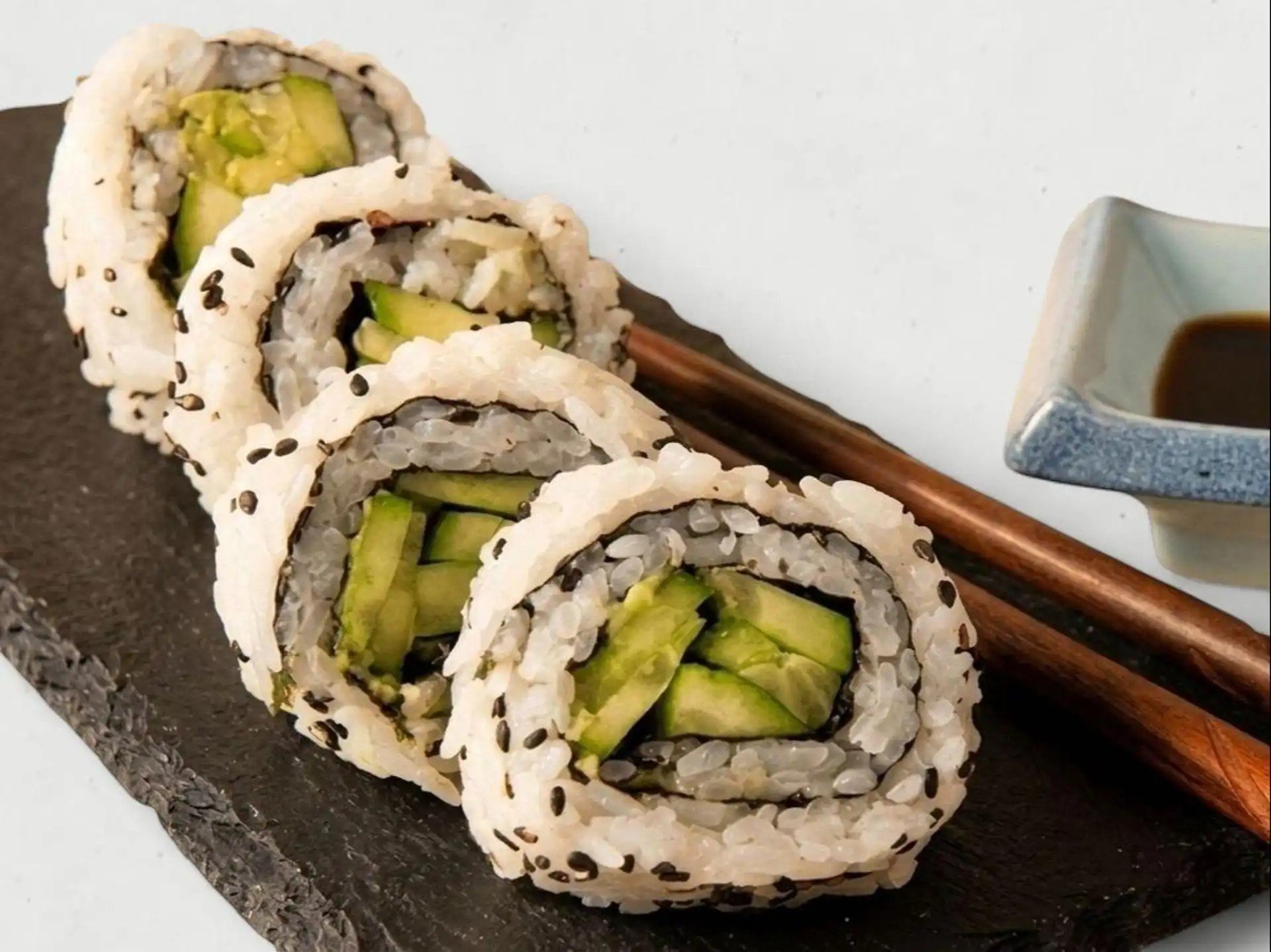 Close-up of sushi rolls with cucumber and rice, garnished with black sesame seeds, on a dark plate with chopsticks and dipping sauce in a small ceramic dish.