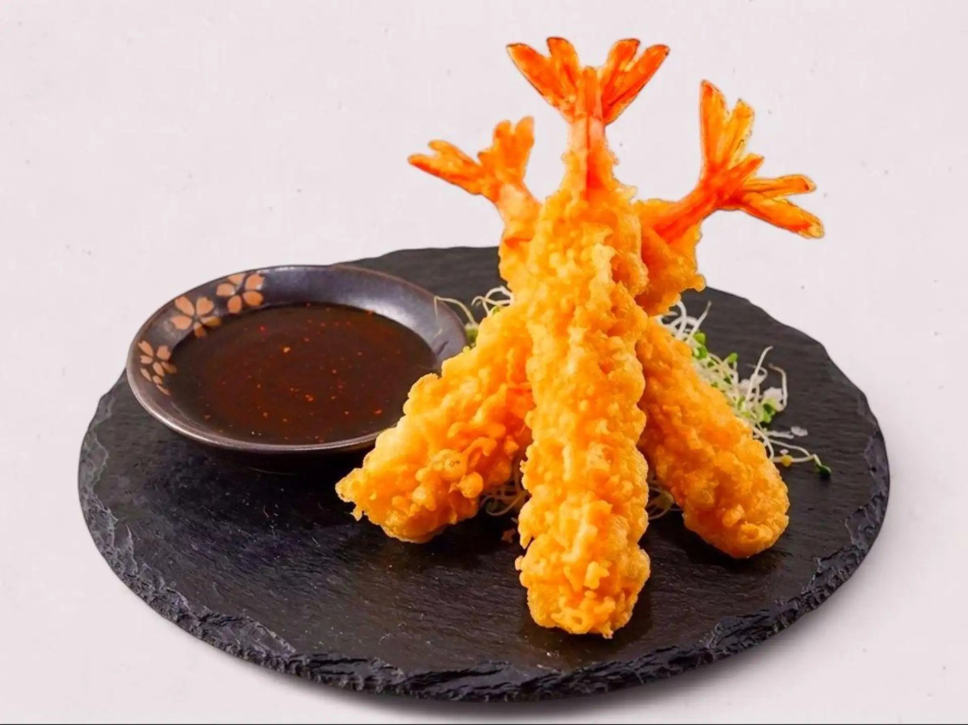 Three pieces of fried shrimp tempura with a small bowl of dipping sauce on a black slate serving platter.