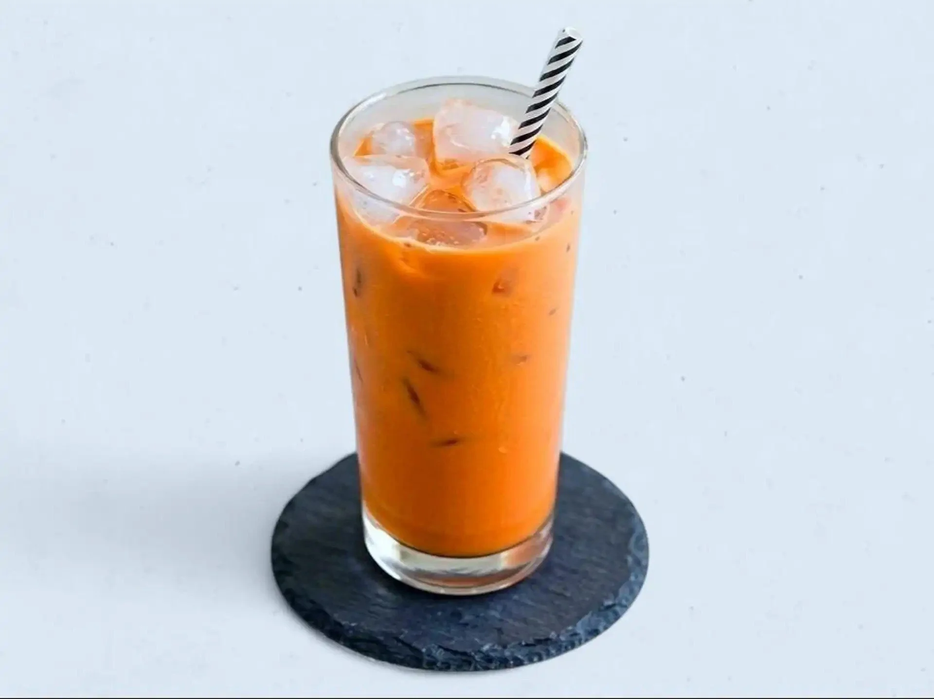 A tall glass of orange-colored iced tea with ice cubes and a striped straw, placed on a round black coaster on a white surface.