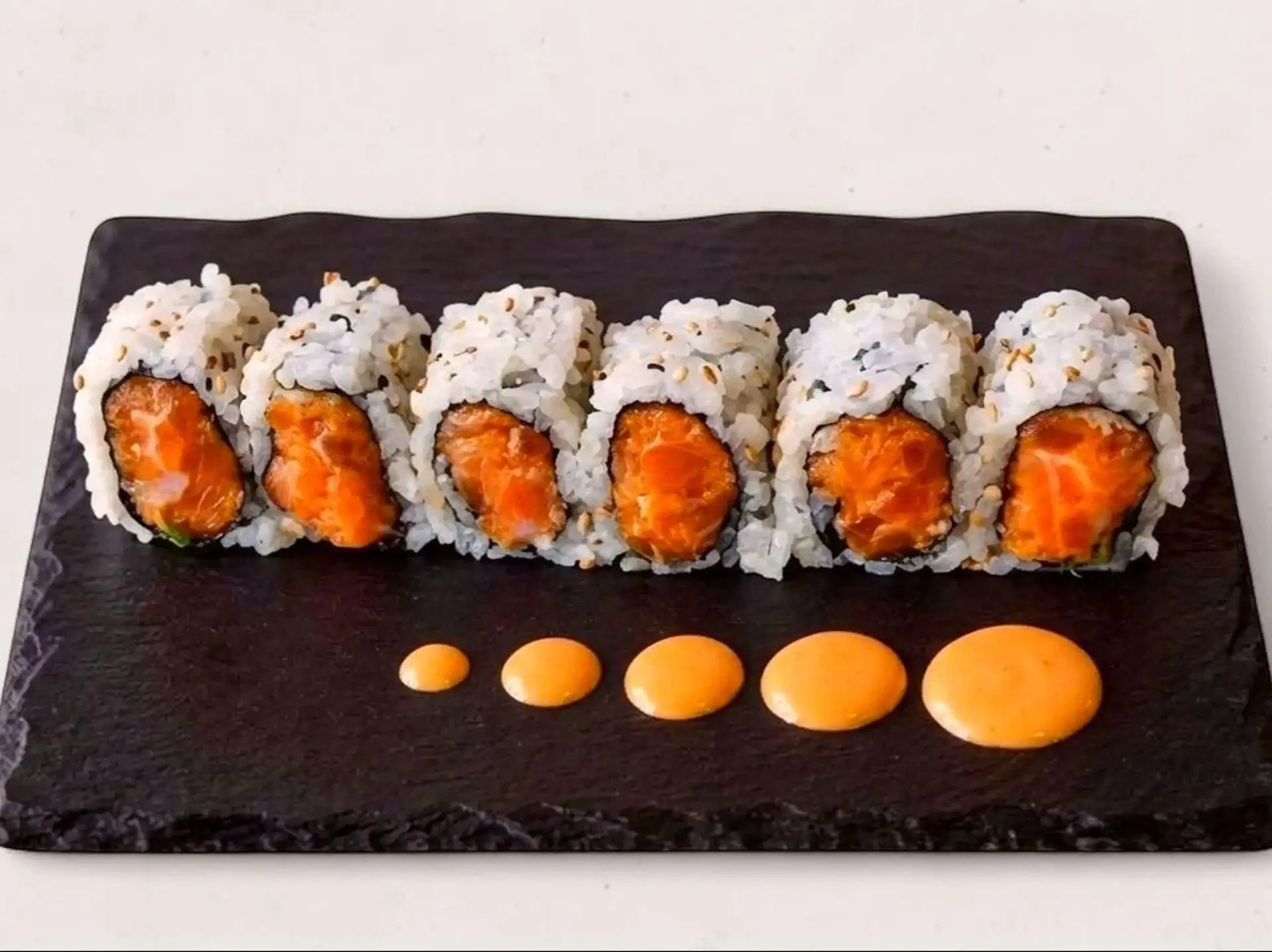 Close-up of five salmon sushi rolls on a black rectangular plate, with a line of yellow and orange candies in front.