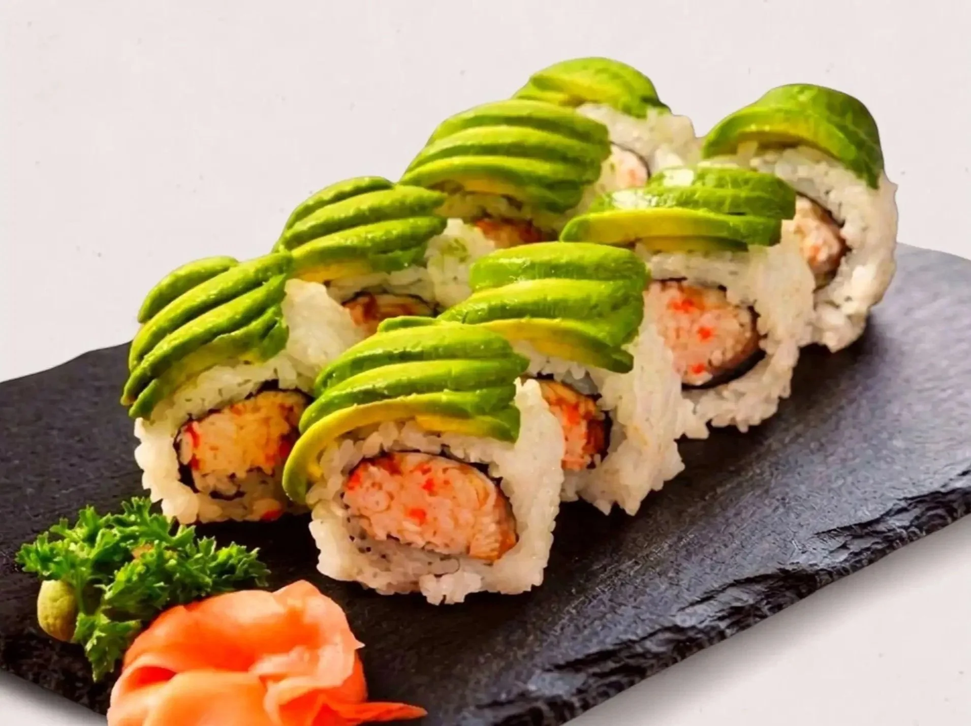 A row of sushi rolls with avocado slices on top, served on a black slate platter with garnishes of parsley and pickled ginger.