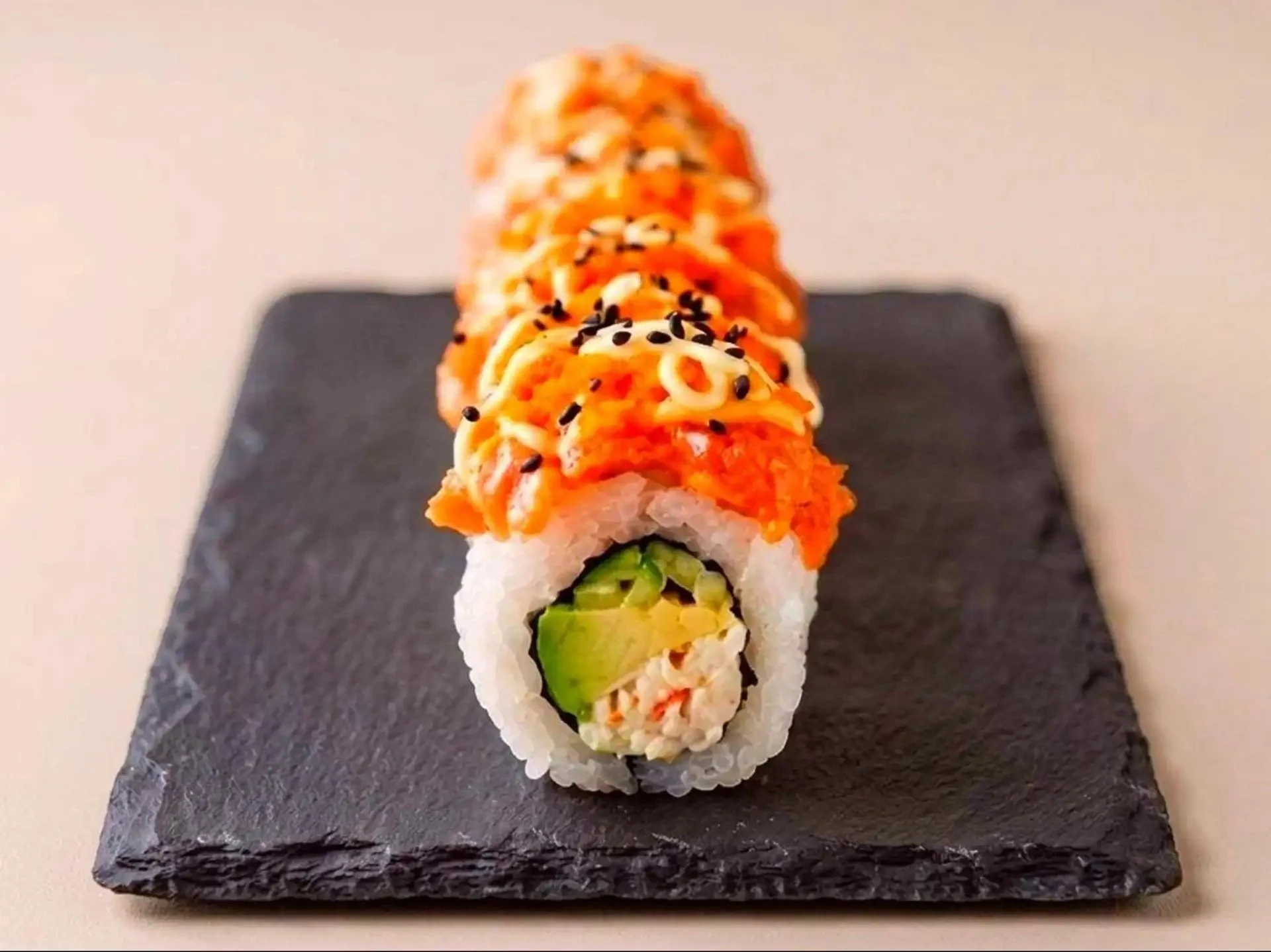 Close-up of a sushi roll on a black slate serving board, showing rice, cucumber, and crab meat inside, topped with orange fish roe, black sesame seeds, and a drizzle of white sauce.