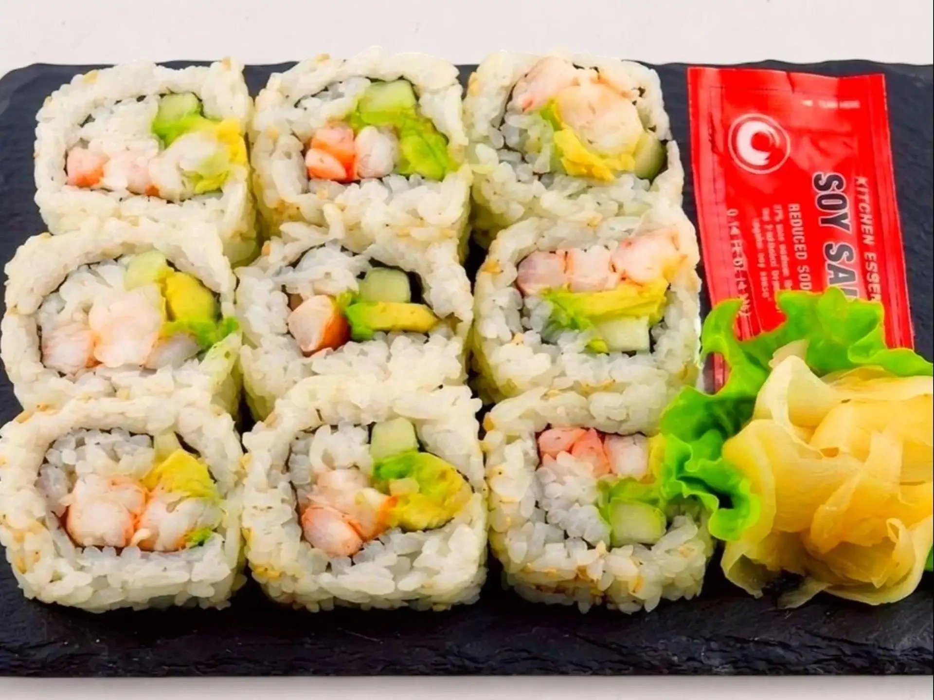 Close-up view of assorted sushi rolls on a black slate platter, topped with seafood, avocado, and cucumber, with a side of salad and a red packet of soy sauce.