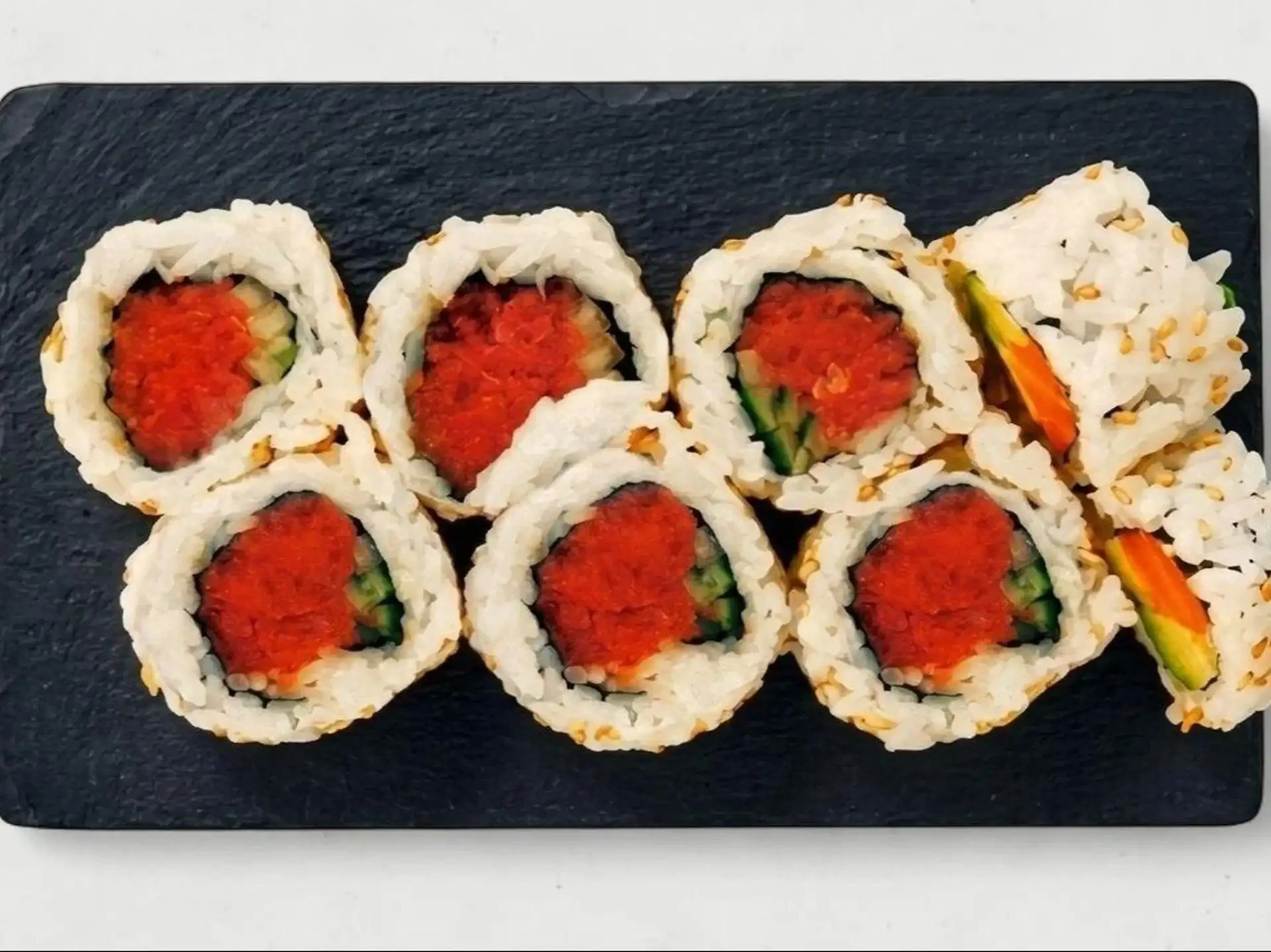 Set of eight sushi rolls on black rectangular plate, filled with rice, tuna, cucumber, and topped with sesame seeds.