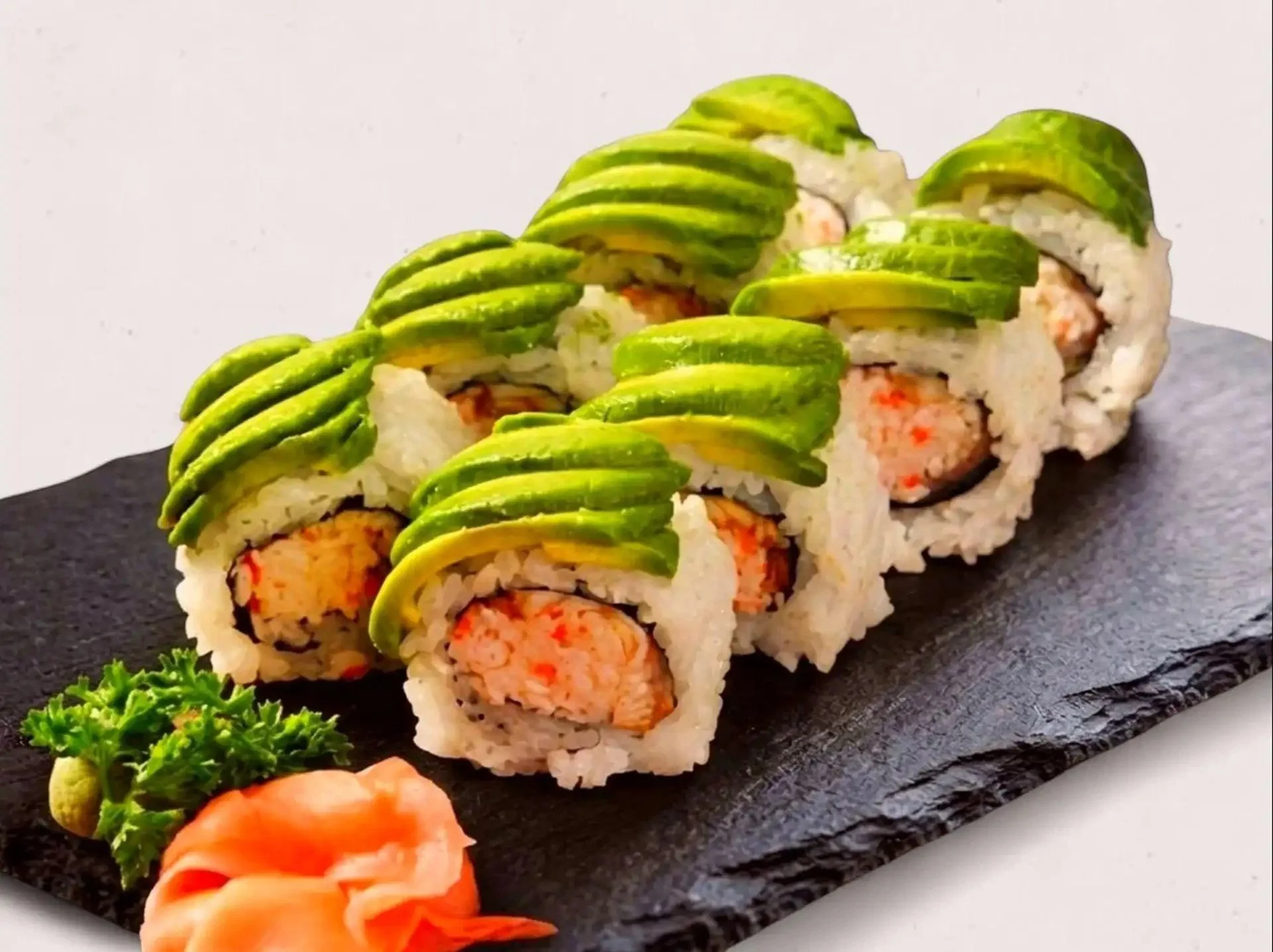 Close-up of a sushi roll topped with thinly sliced avocado on a black slate serving platter, garnished with parsley and pickled ginger.