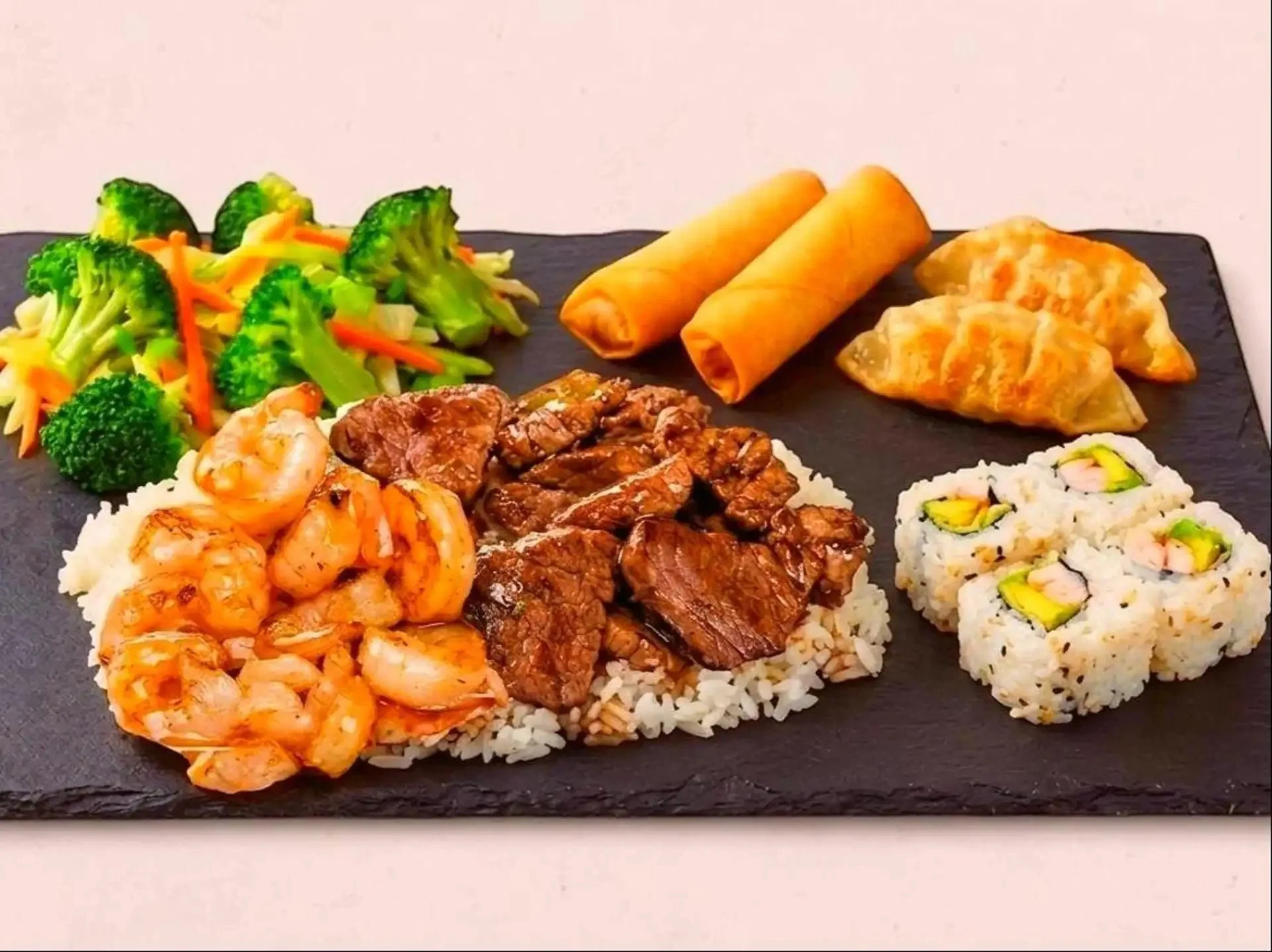 Assorted sushi rolls and Asian dishes on a black slate serving platter, including fried spring rolls, shrimp and seafood, beef and chicken over rice, and a small side of pickled ginger on a white background.