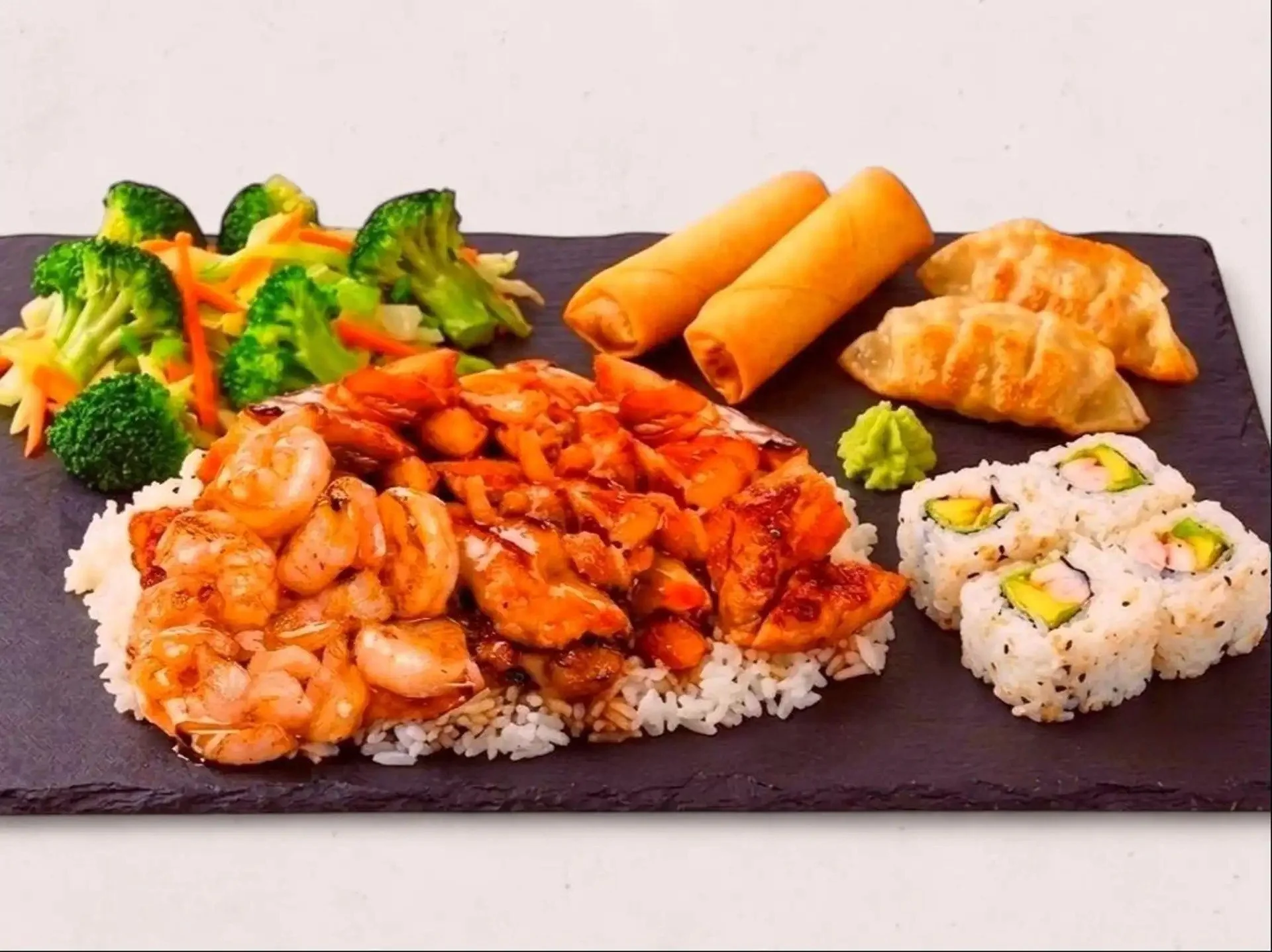 Assorted sushi rolls, shrimp, fried spring rolls, and a vegetable stir-fry with broccoli and carrots served on a black rectangular plate.