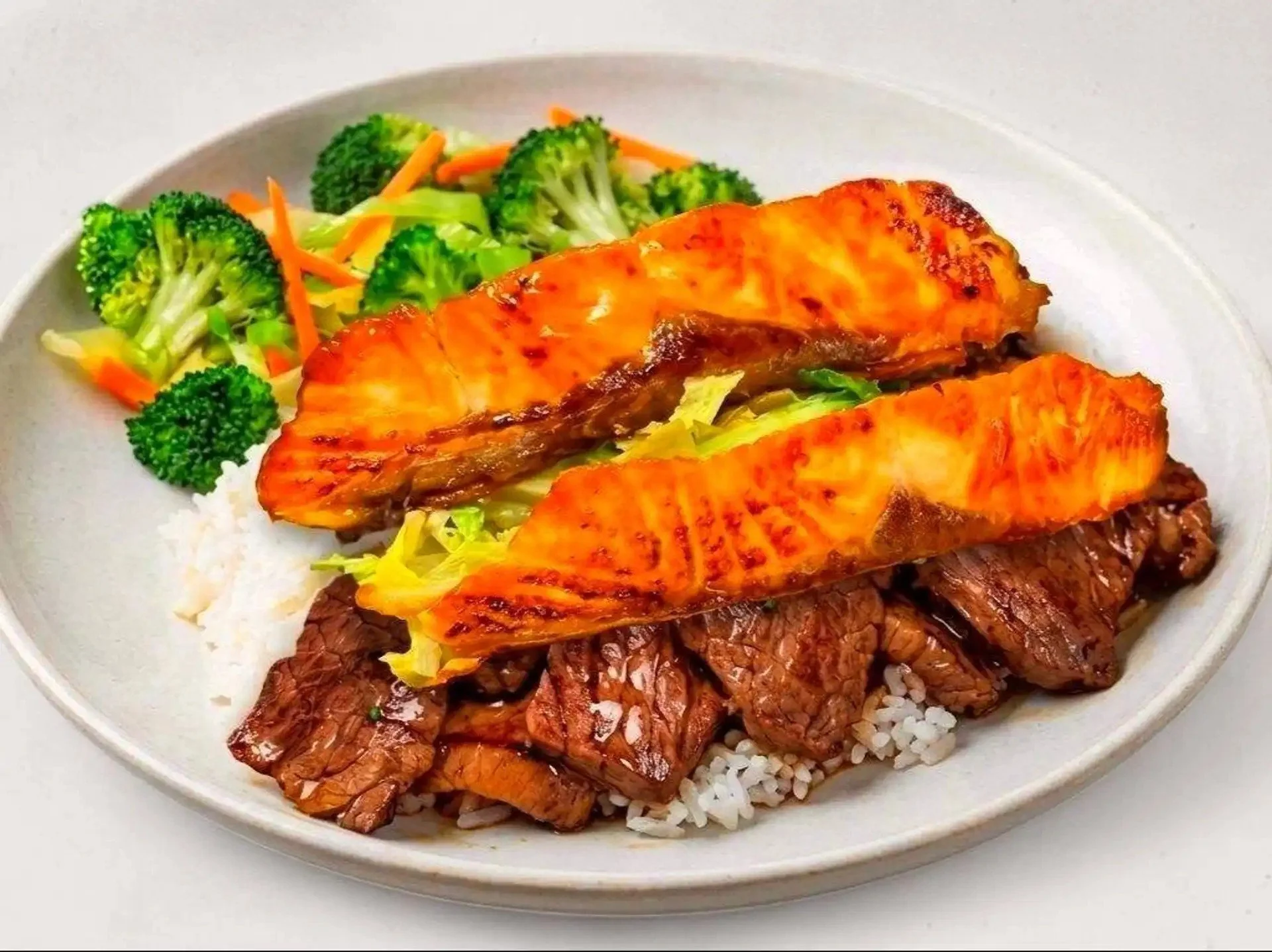 A plate of rice topped with grilled meat, grilled salmon, and a side of steamed broccoli and shredded carrots.