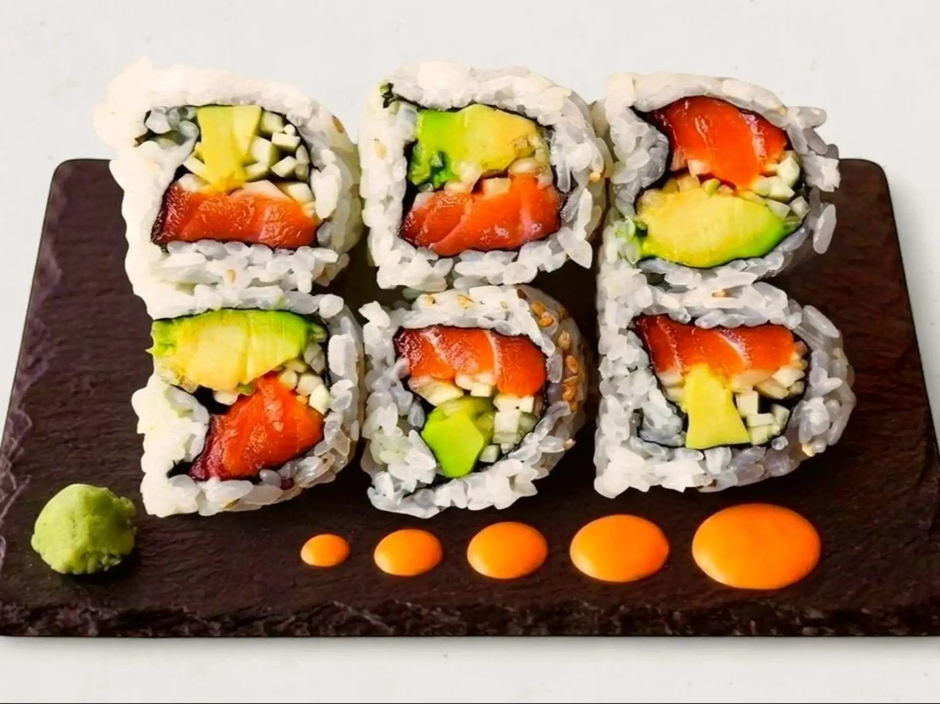 Close-up of six pieces of sushi rolls with rice and fresh fish and vegetables, served on a dark wooden platter with small dollops of orange sauce and wasabi around it.