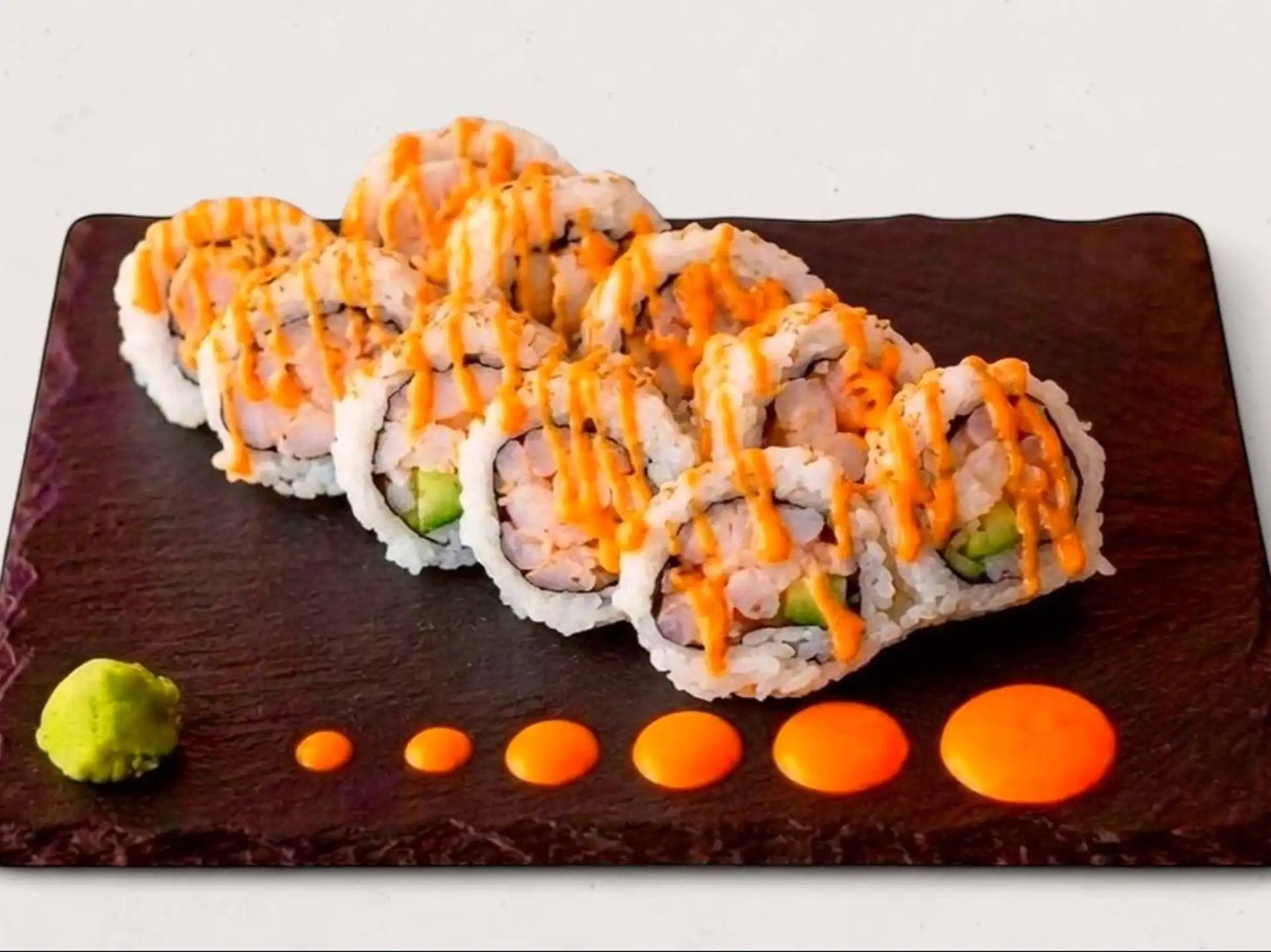 Sushi rolls topped with orange sauce on a dark rectangular plate, garnished with wasabi and pickled ginger on a white background.