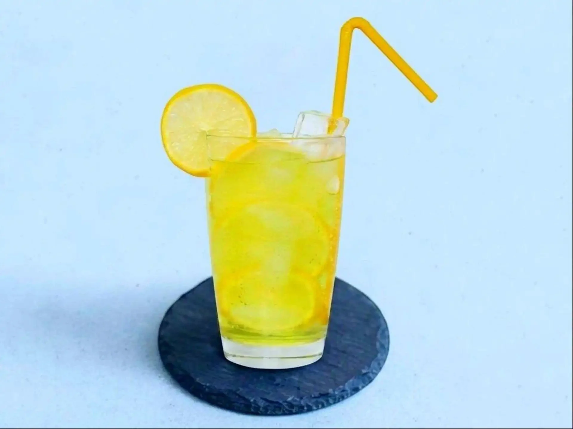 A glass of lemonade with a slice of lemon and ice cubes, a yellow straw, sitting on a dark coaster on a light background.