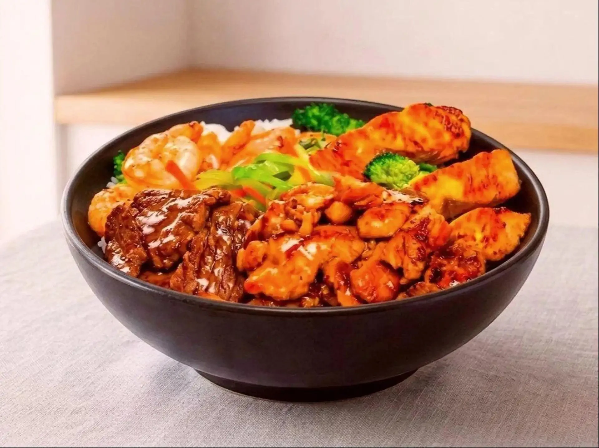 Black bowl filled with rice, fried chicken, broccoli, and various stir-fried vegetables and meat in a sauce, placed on a white surface with a wooden backdrop.