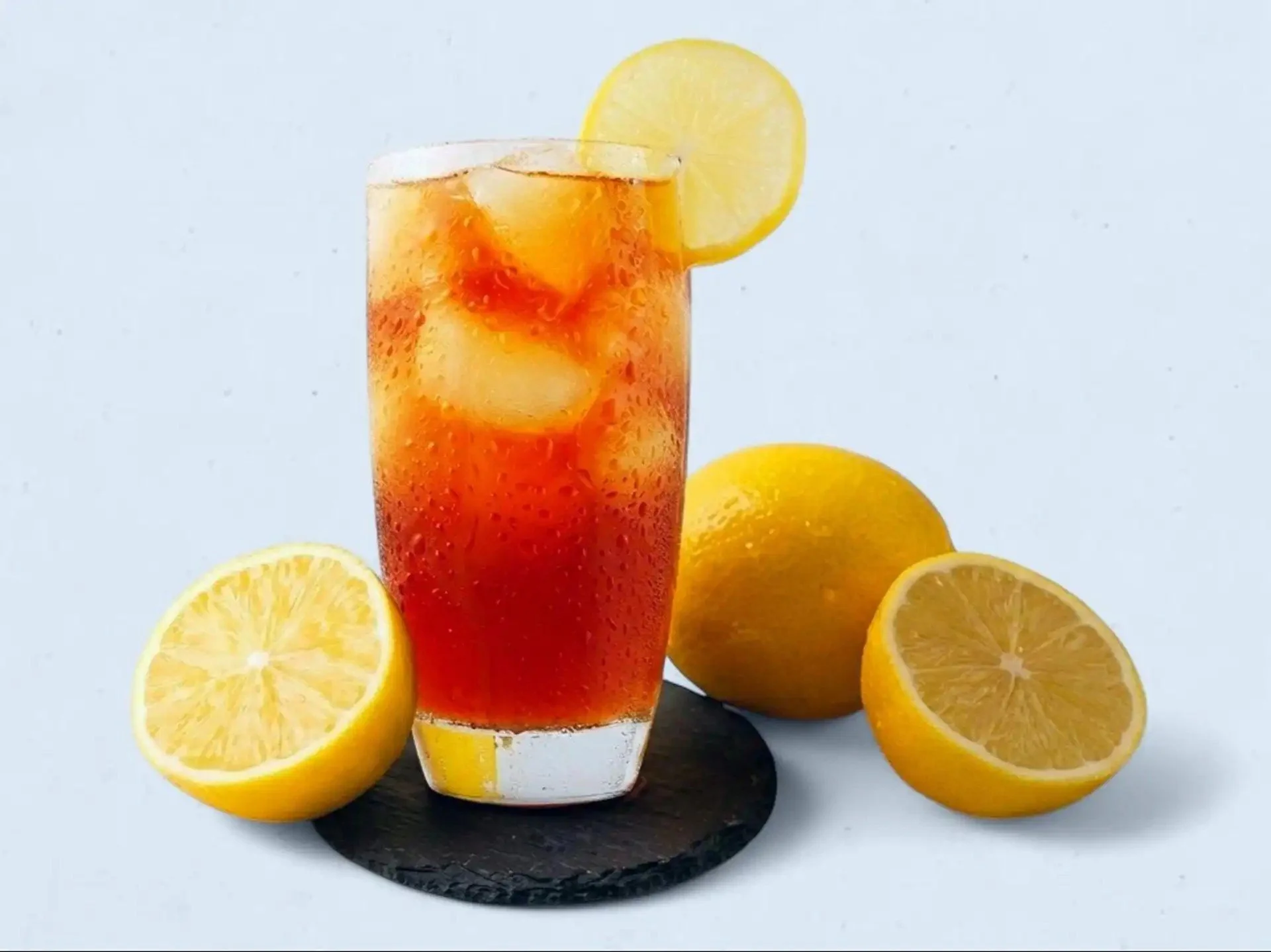 A glass of iced tea with lemon slices and whole lemons on a black slate. The glass has a lemon slice garnish and ice cubes inside.