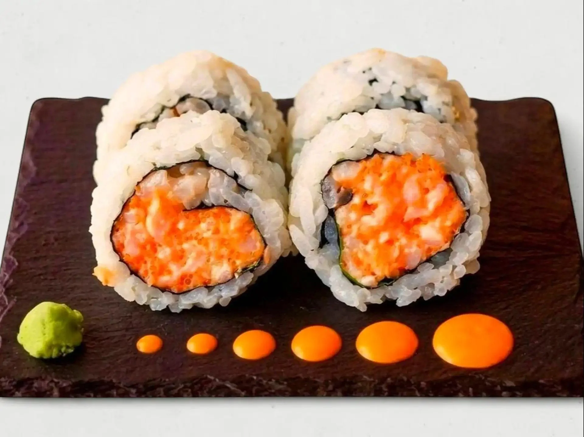 Close-up of four sushi rolls on a black plate, filled with orange fish and wrapped in white rice and seaweed, garnished with green wasabi and small orange sauce dots.