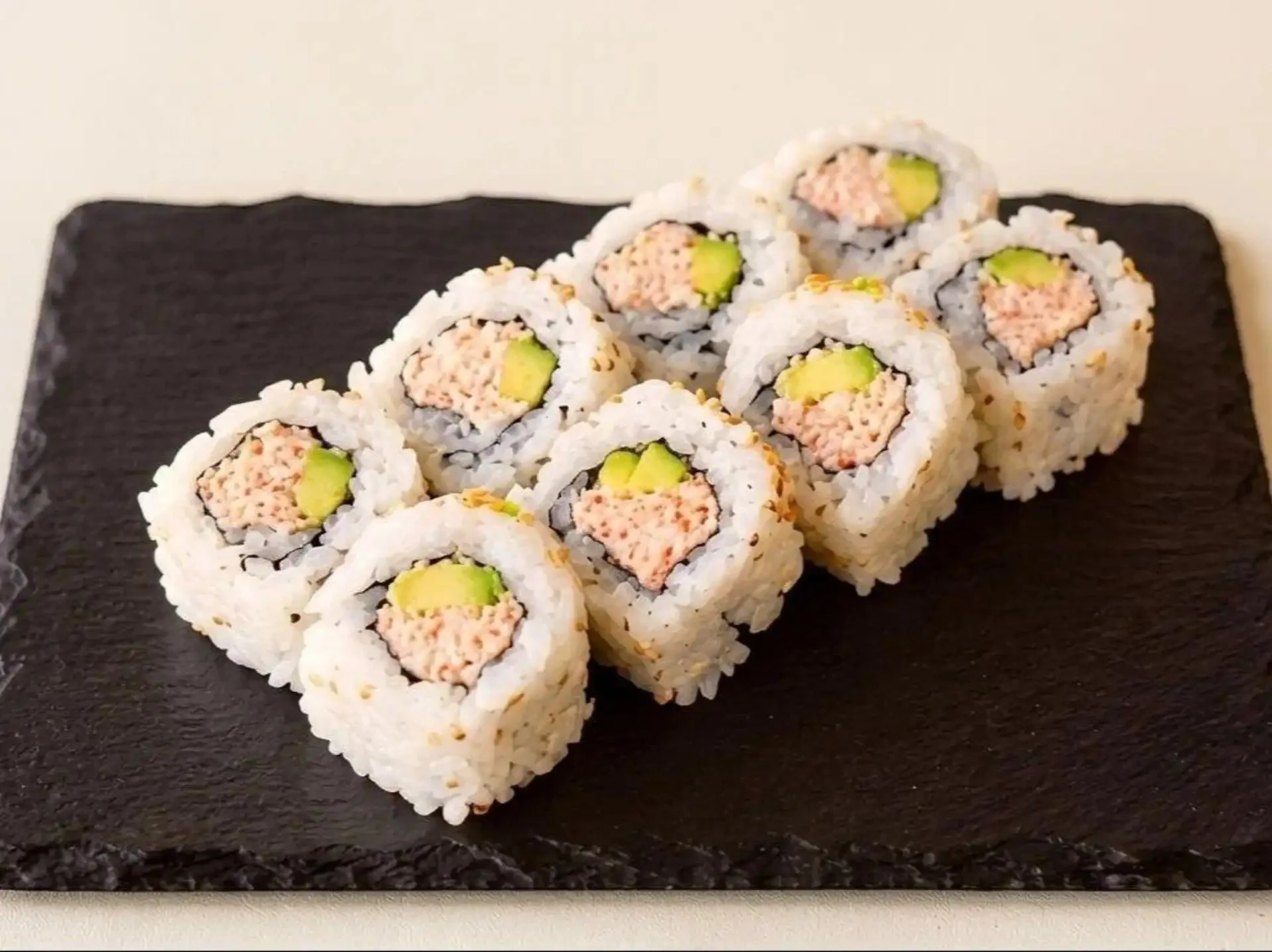 Close-up of six sushi rolls with rice, avocado, and cooked fish, arranged on a black slate serving platter.