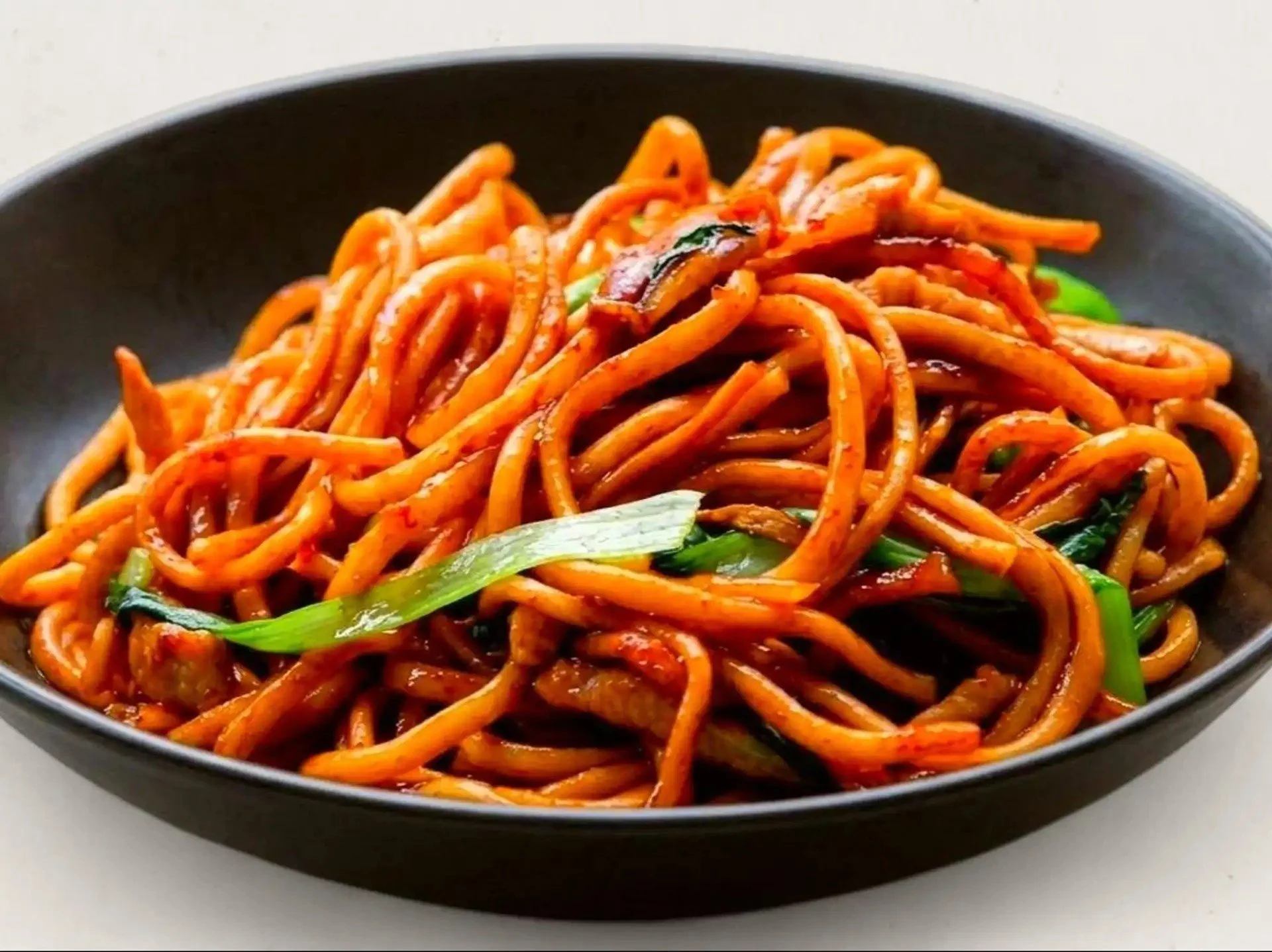 A black bowl filled with Asian-style stir-fried noodles in a savory sauce, garnished with green onions.