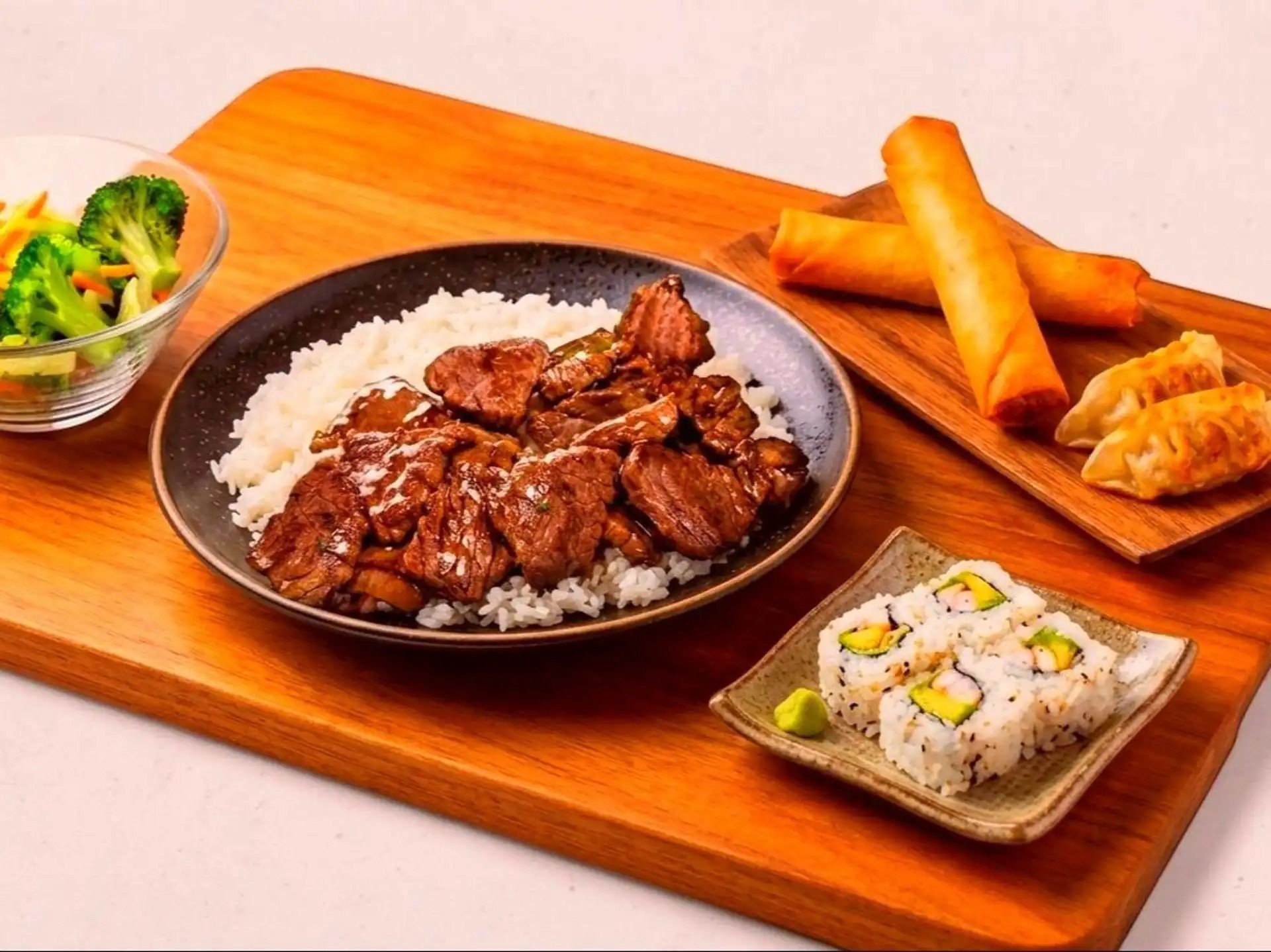 Assorted Japanese dishes on a wooden serving board including beef with rice, spring rolls, sushi rolls, and salad.