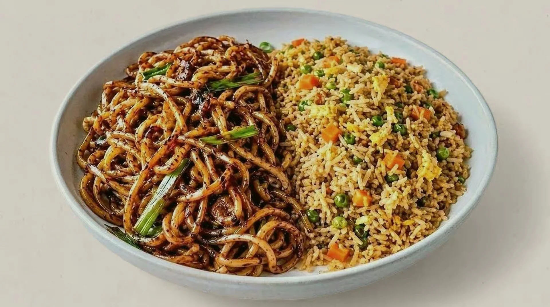 A bowl containing a portion of Chinese chow mein noodles garnished with green onions on the left side, and a serving of fried rice mixed with peas and diced carrots on the right side.